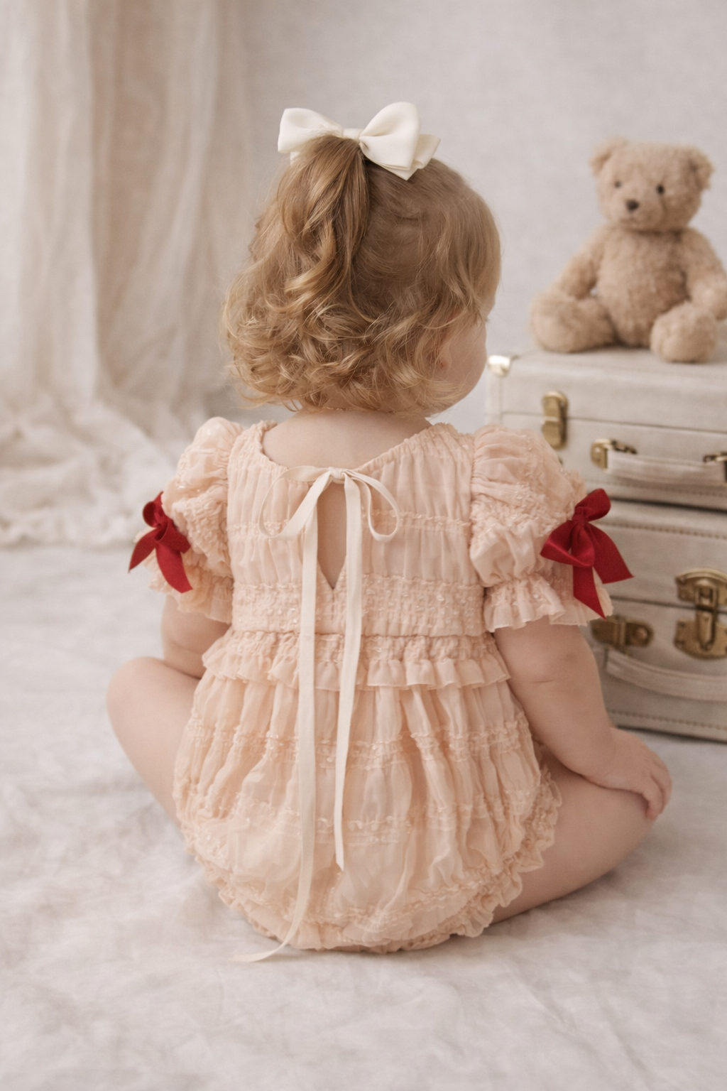Child in a lace dress with red bows sitting on the floor next to a teddy bear and suitcase.