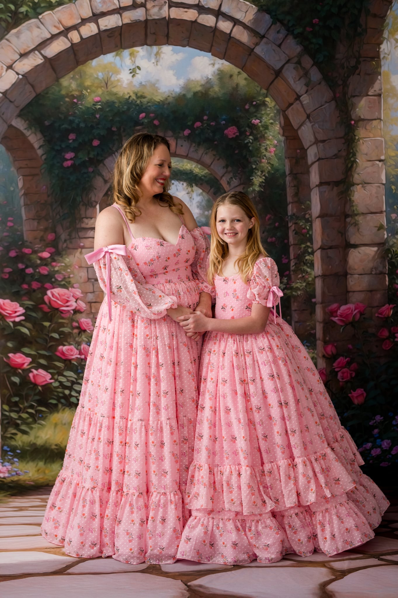 Two women in pink dresses standing under a stone archway with floral decorations.