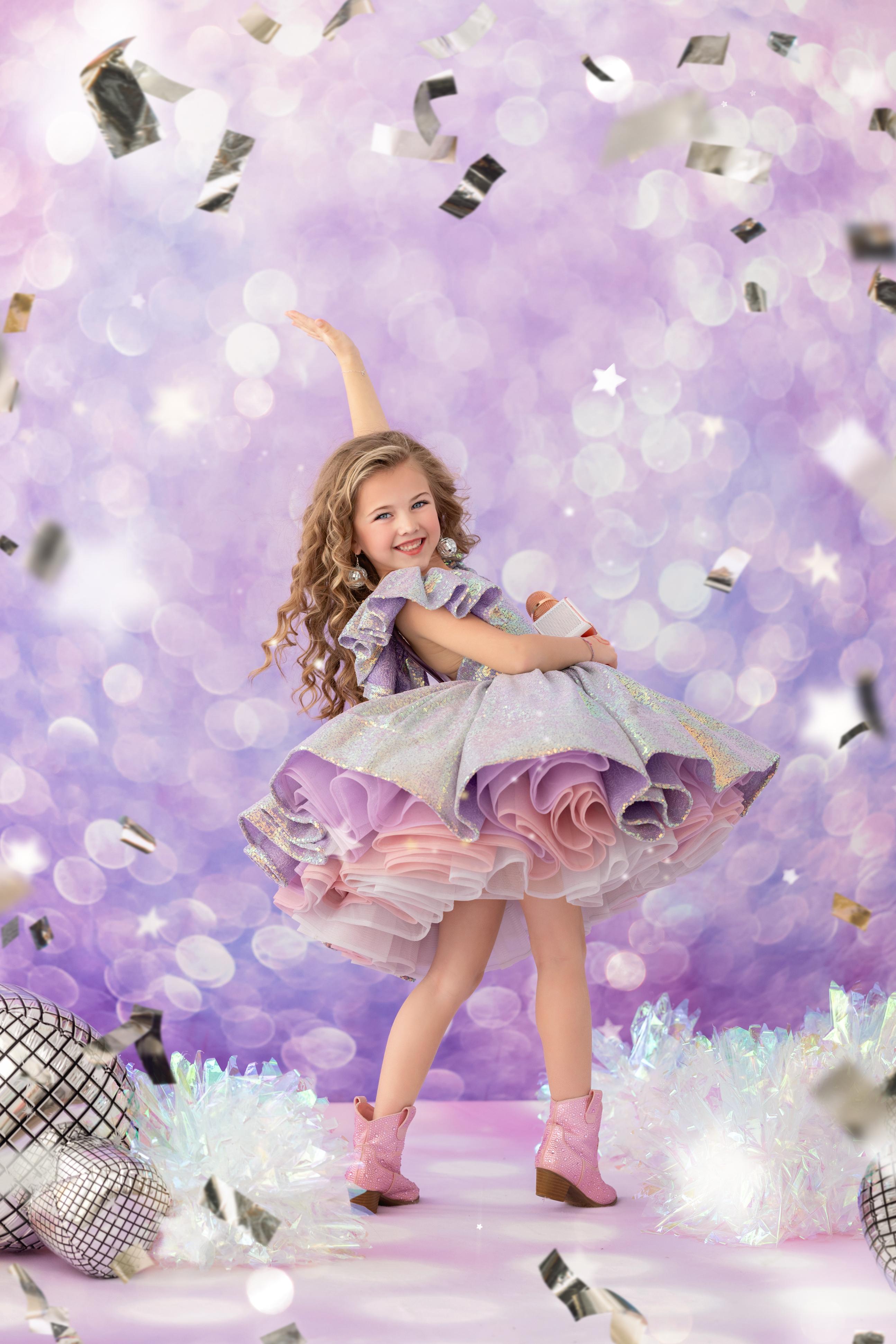 A young girl wearing a lilac-colored gown with a tulle skirt, standing in a pose that showcases the dress, with confetti falling around her.