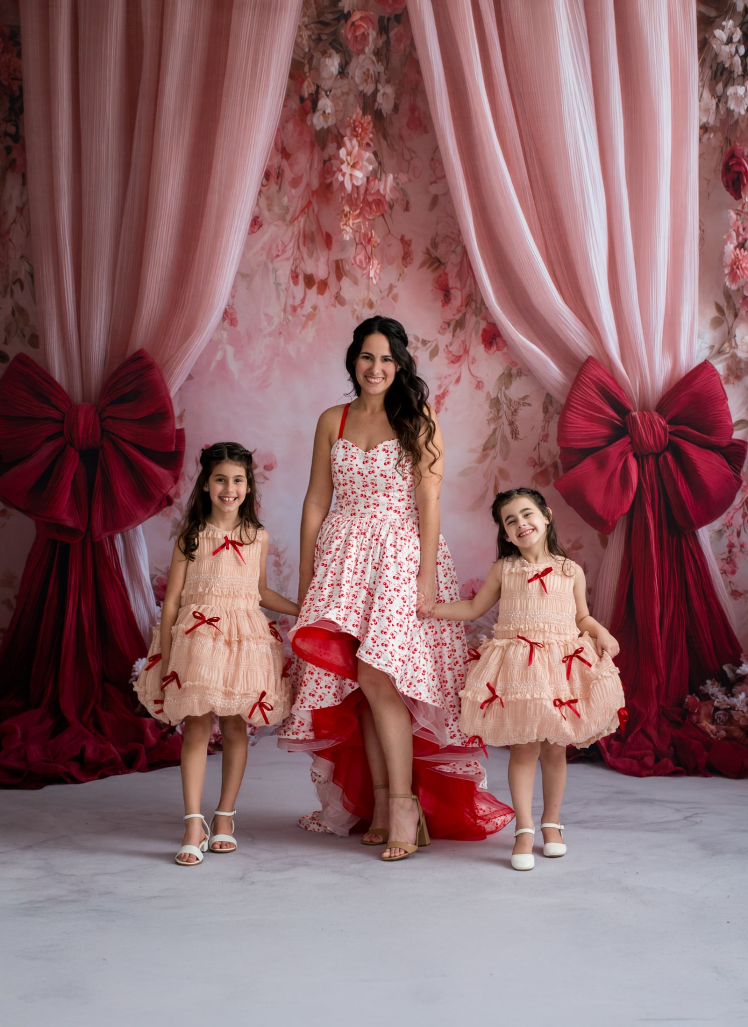 Woman with two young girls in matching dresses standing in front of pink floral curtains with red bows.