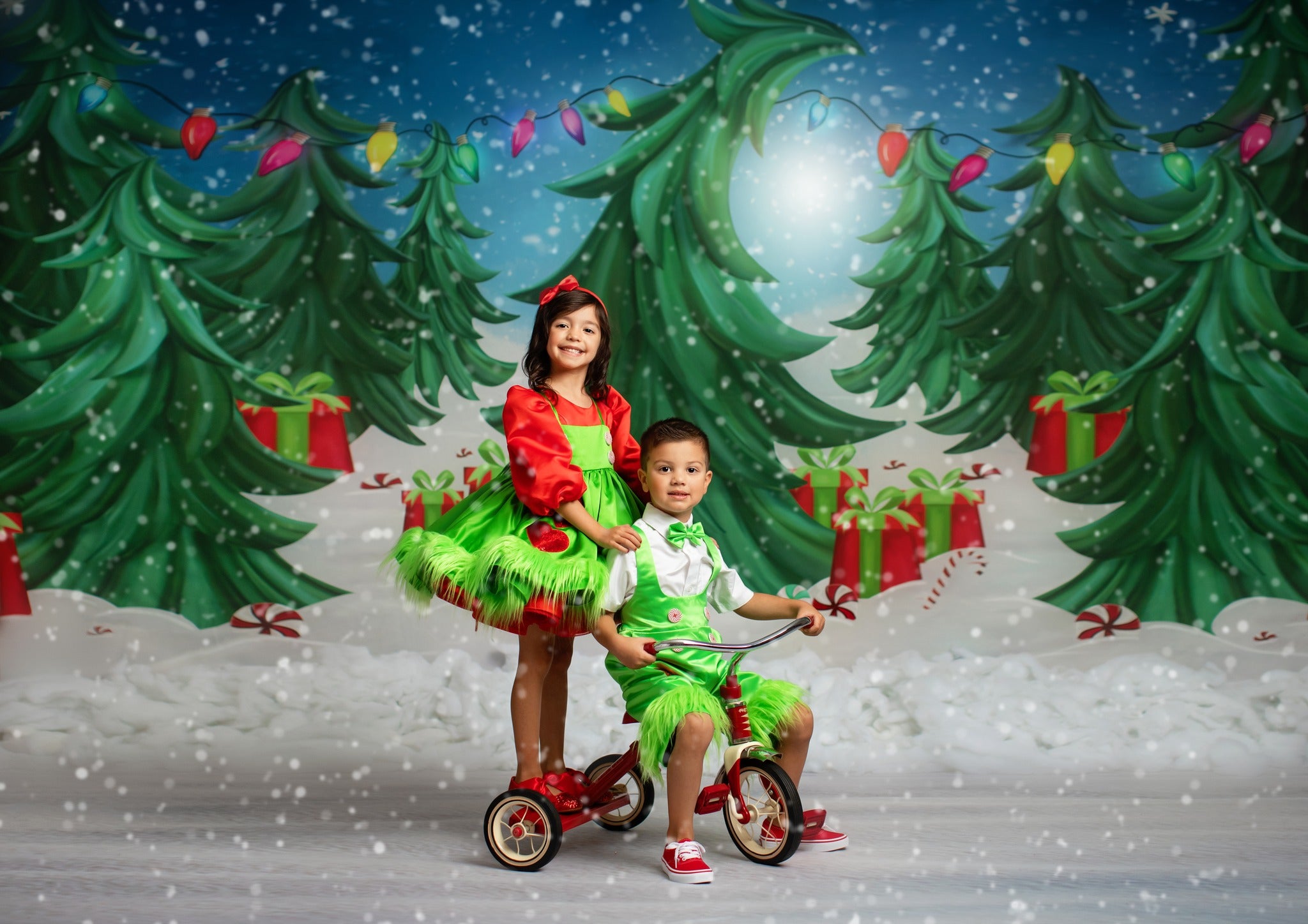 Two children dressed in vibrant green and red Christmas-themed outfits, smiling in front of a festive holiday backdrop with decorated trees and gifts.