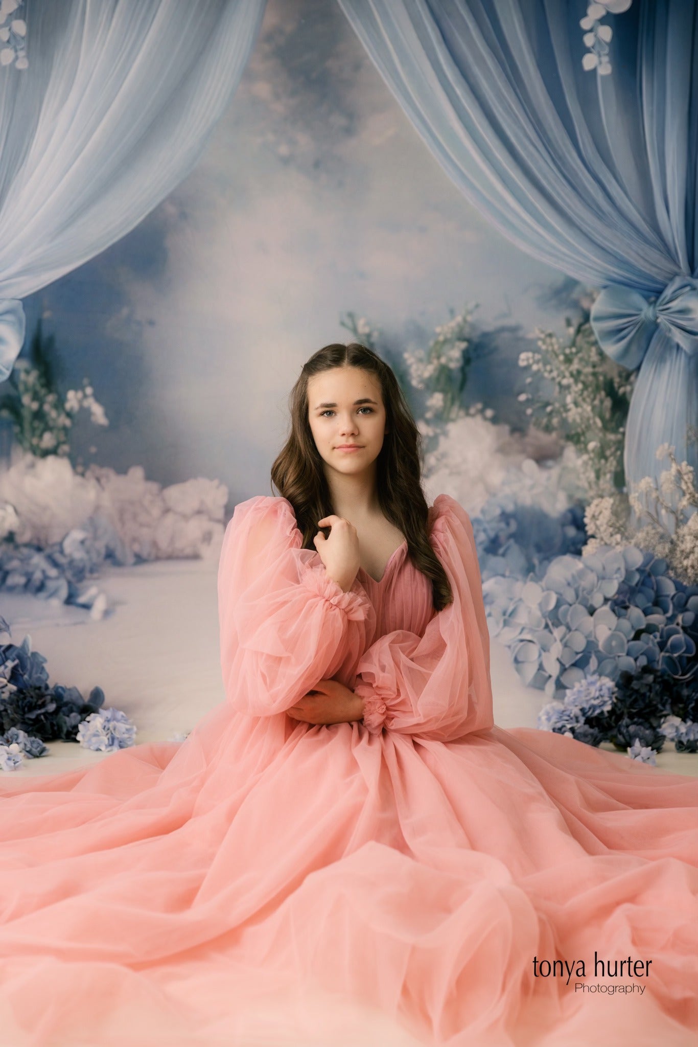 A girl in a voluminous pink gown stands against an ethereal blue backdrop decorated with floral arrangements, bows, and flowing curtains.