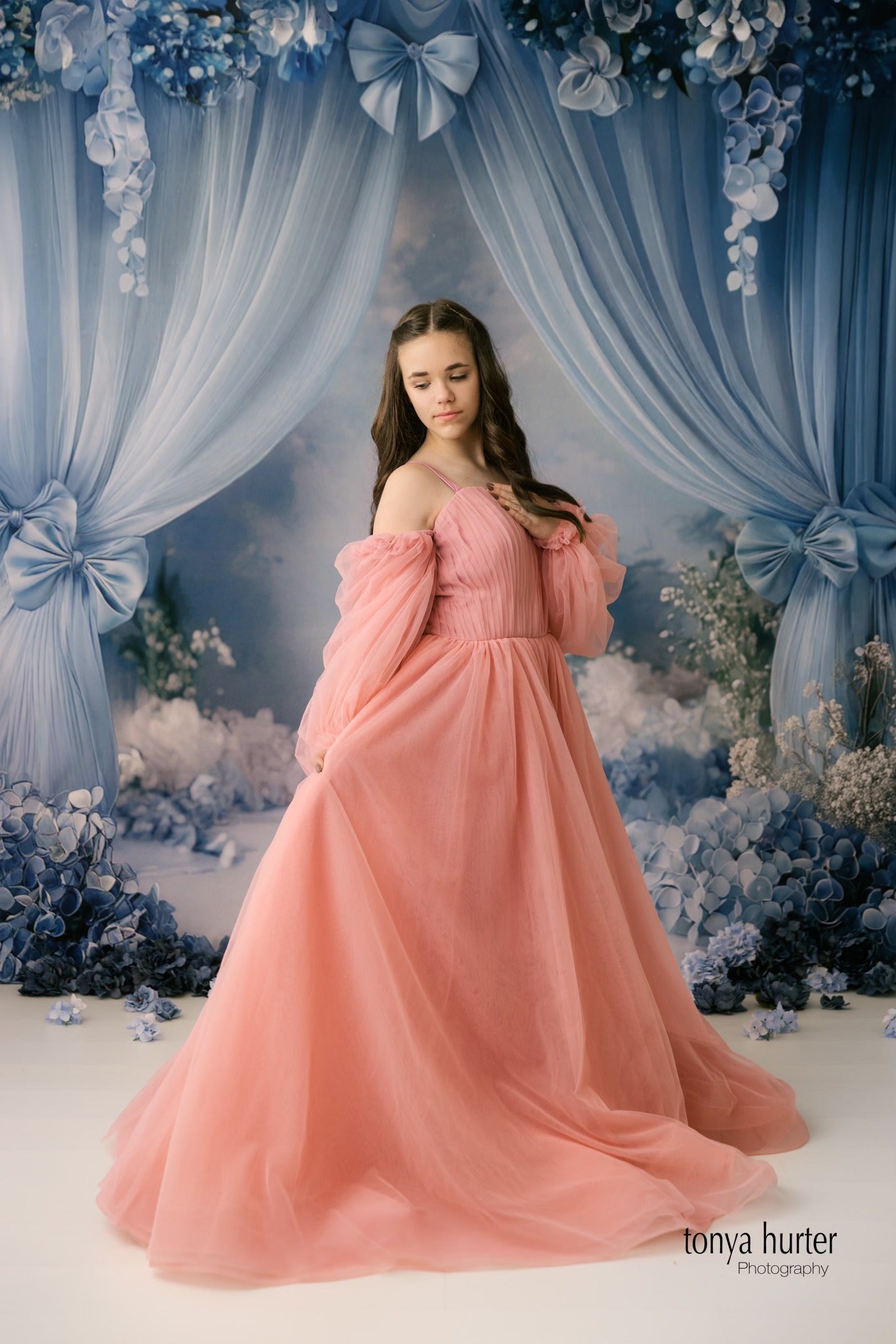 A soft and romantic portrait of a girl in a pink gown, gracefully holding the skirt, set against a blue floral scene with cascading fabric and hydrangea details.