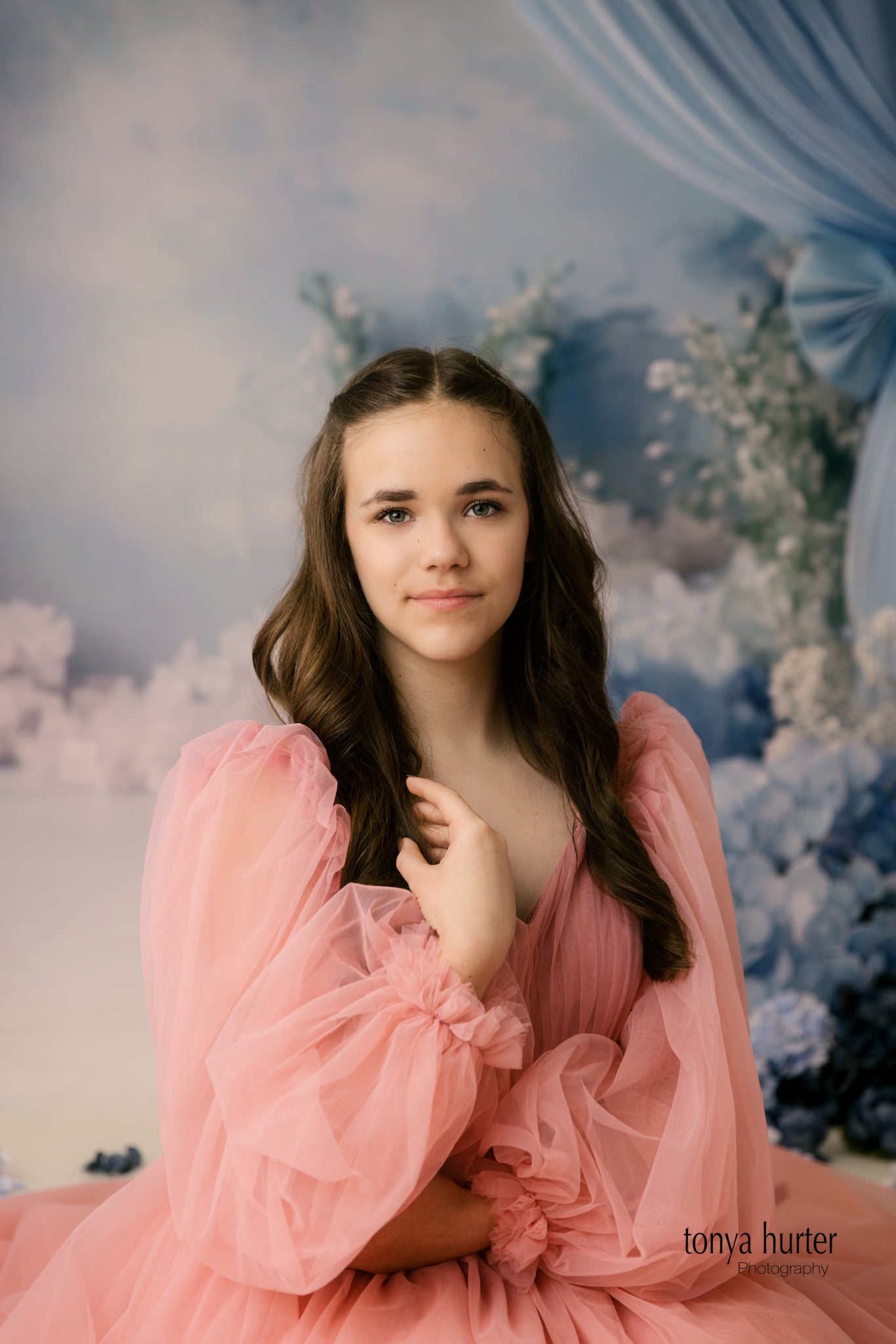 A young woman in a pastel pink off-the-shoulder dress poses with a serene expression in a fantasy-inspired setting featuring blue flowers and sheer drapery.