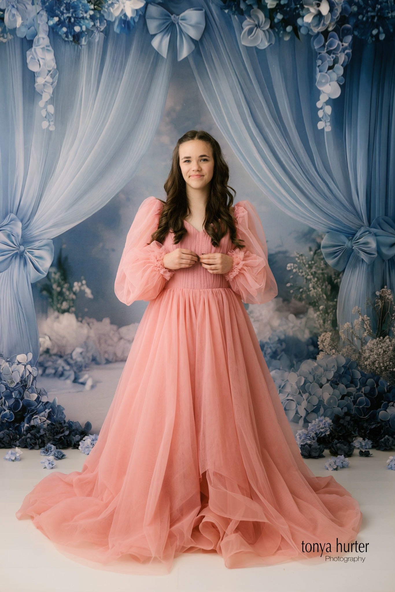 A young girl with long, wavy brown hair wears an elegant flowing pink gown with off-the-shoulder tulle sleeves, standing in front of a dreamy blue floral backdrop with draped fabric and bows.