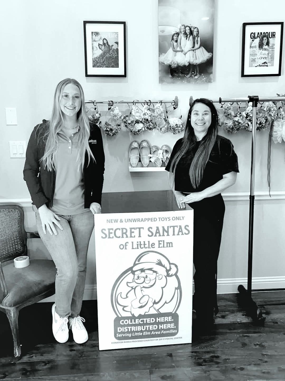 Two women standing next to a 'Secret Santas of Little Elm' sign in a room with decor and framed pictures.