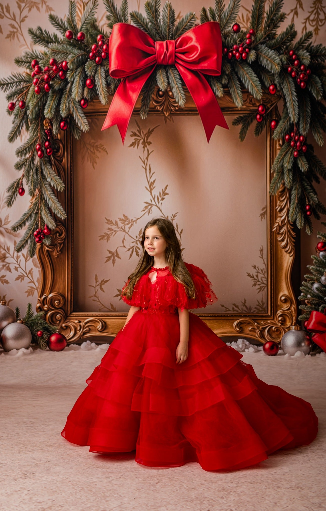 Young girl in a red dress standing in front of a decorated Christmas frame with a large red bow.
