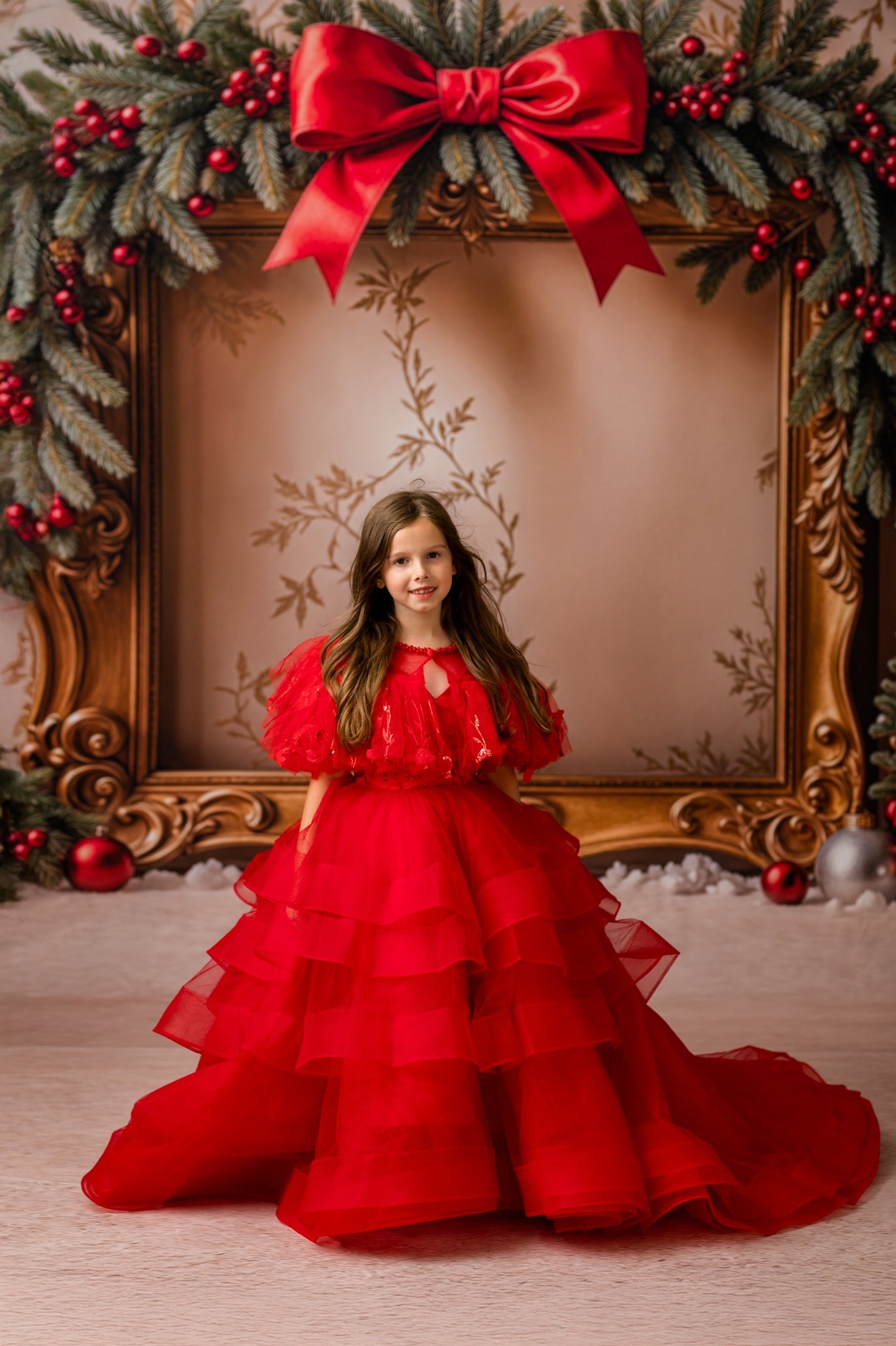 Young girl in a red dress standing in front of a decorated Christmas frame.