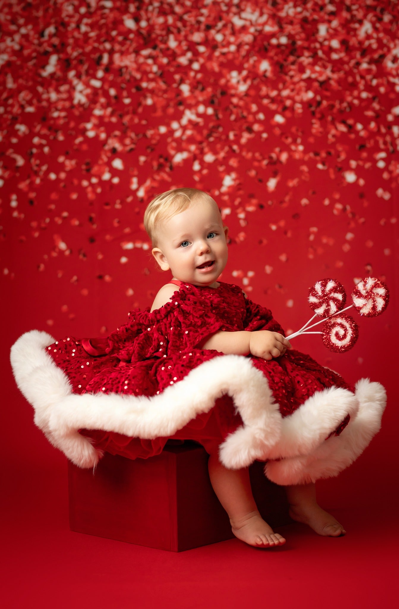 Child in a red dress with white fur trim sitting on a red box against a red background with candy cane decorations.