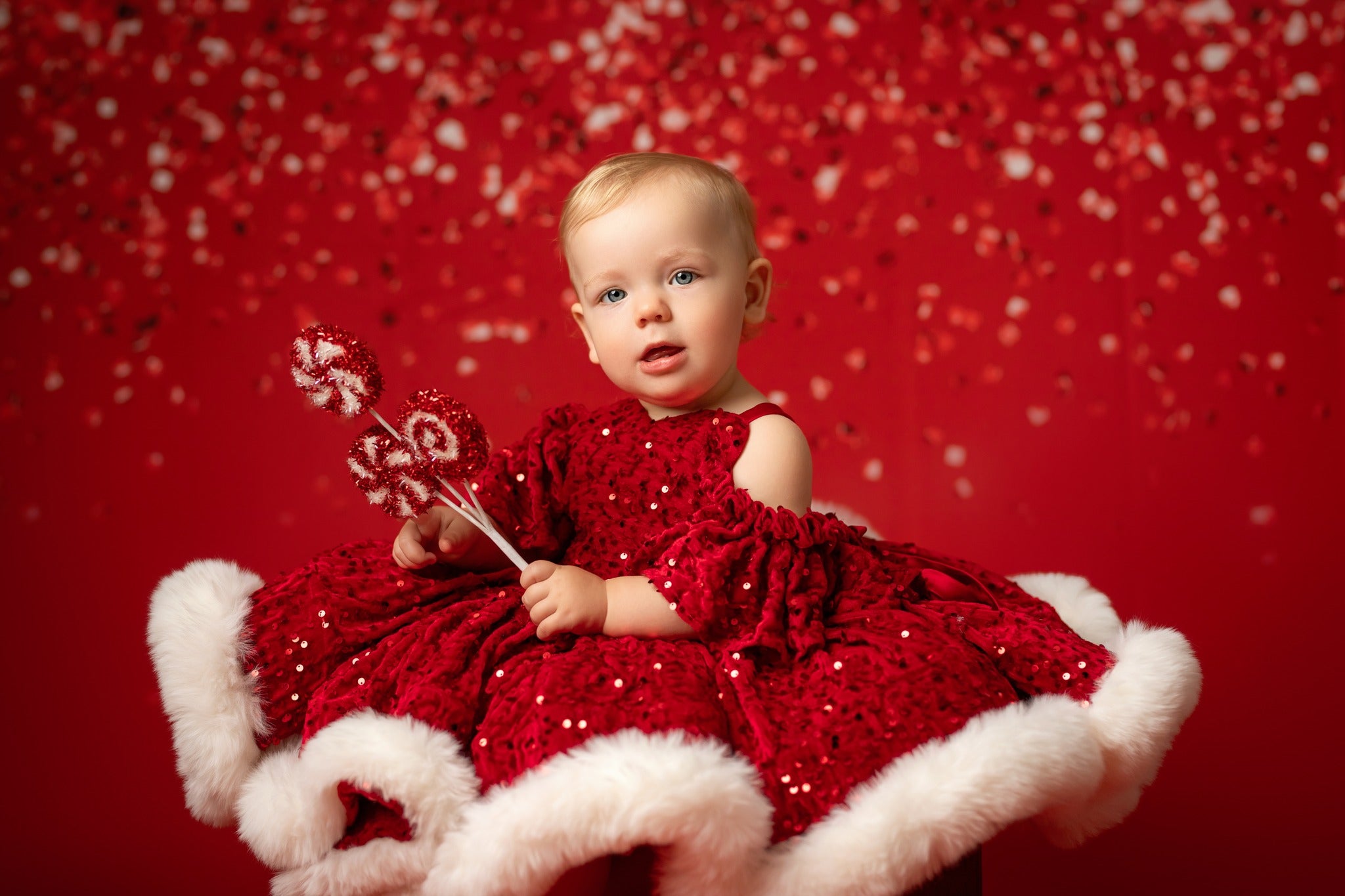 Baby in a red dress with white fur trim sitting on a red background