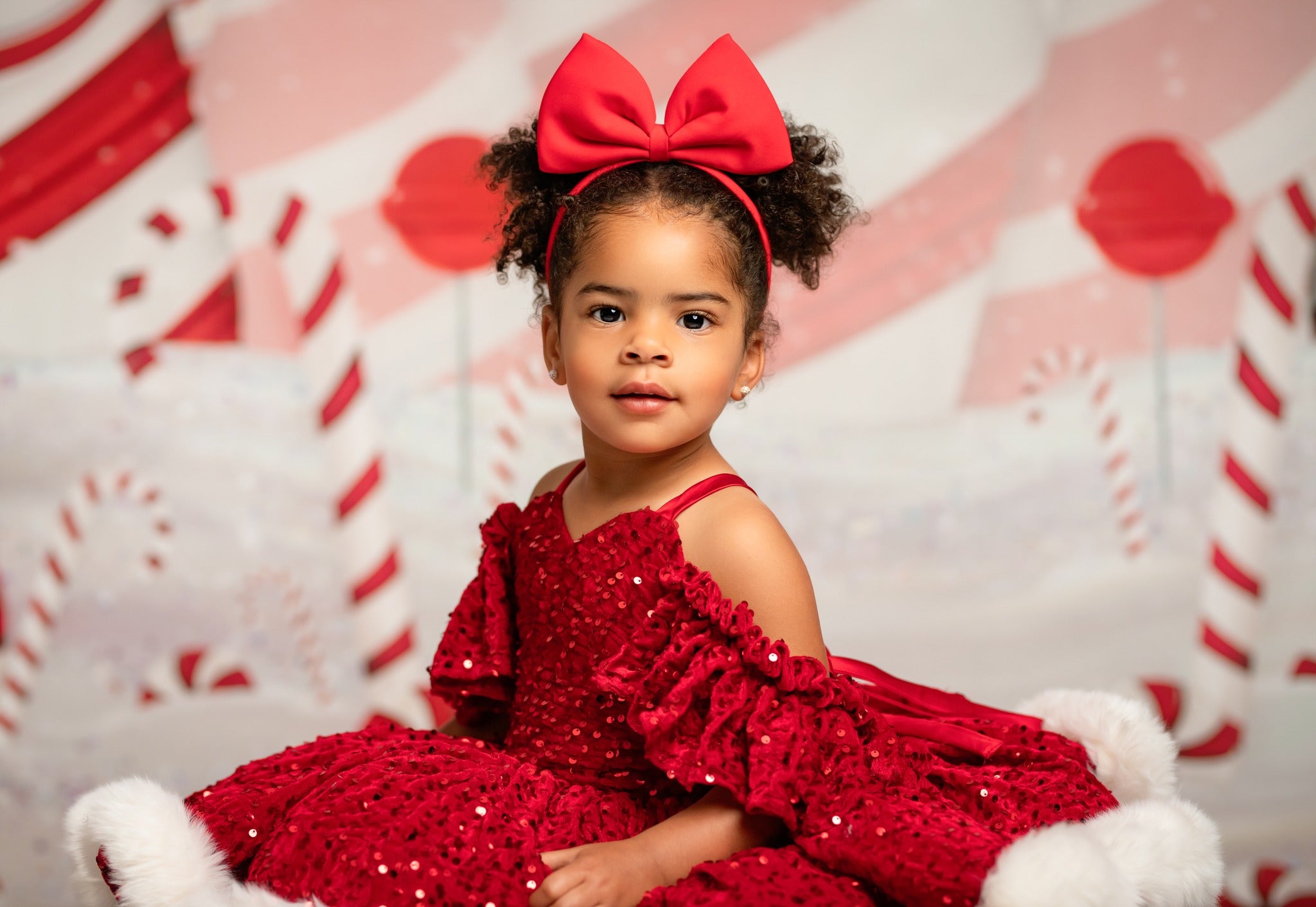 Child in a red sequin dress with a large bow against a candy cane and snowflake patterned background