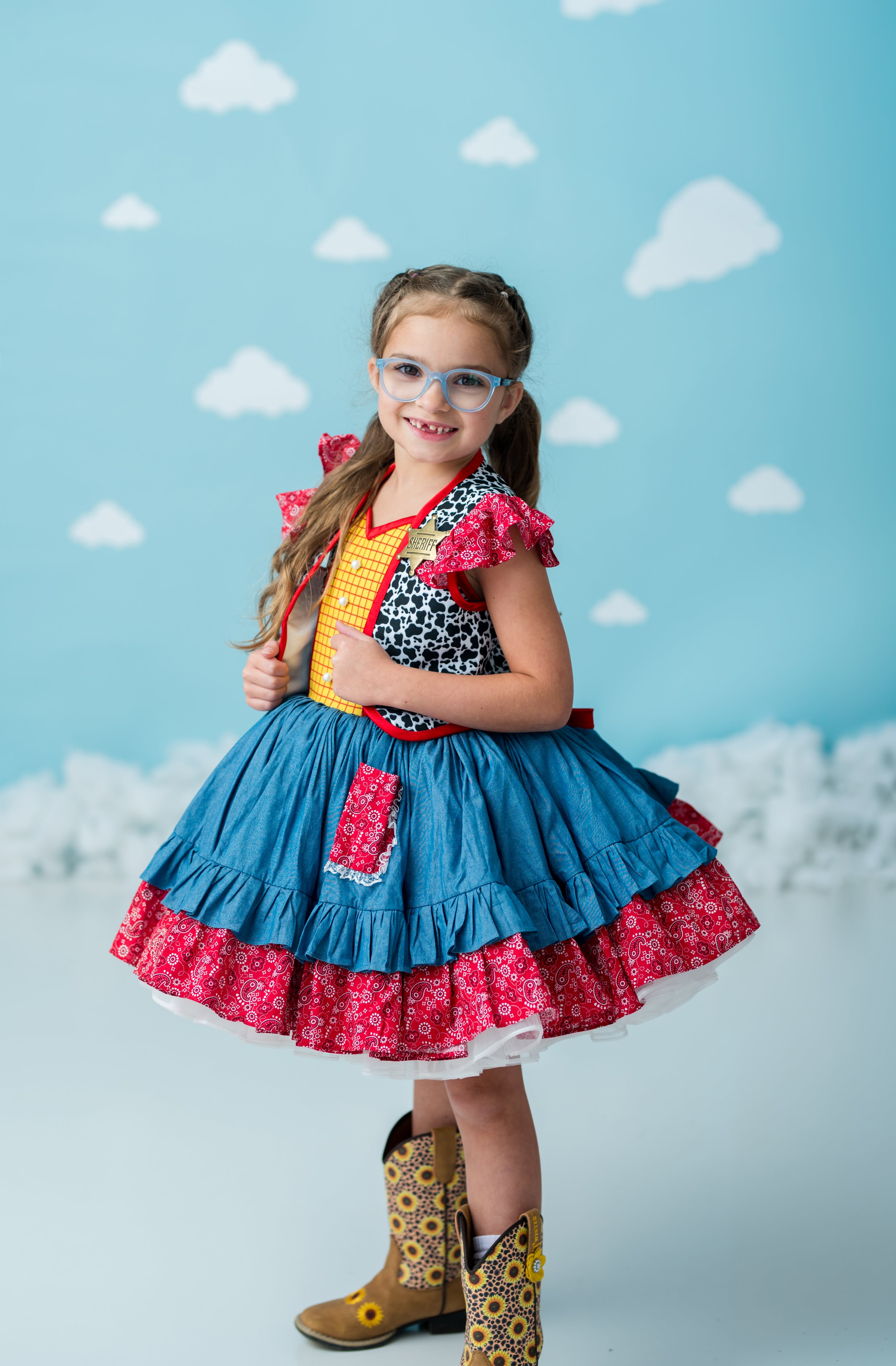 A young girl modeling a western-themed costume dress with a blue tiered skirt, red floral ruffle trim, a yellow vest, and a black-and-white patterned top, paired with cowboy boots, against a blue studio backdrop with white cloud cutouts.