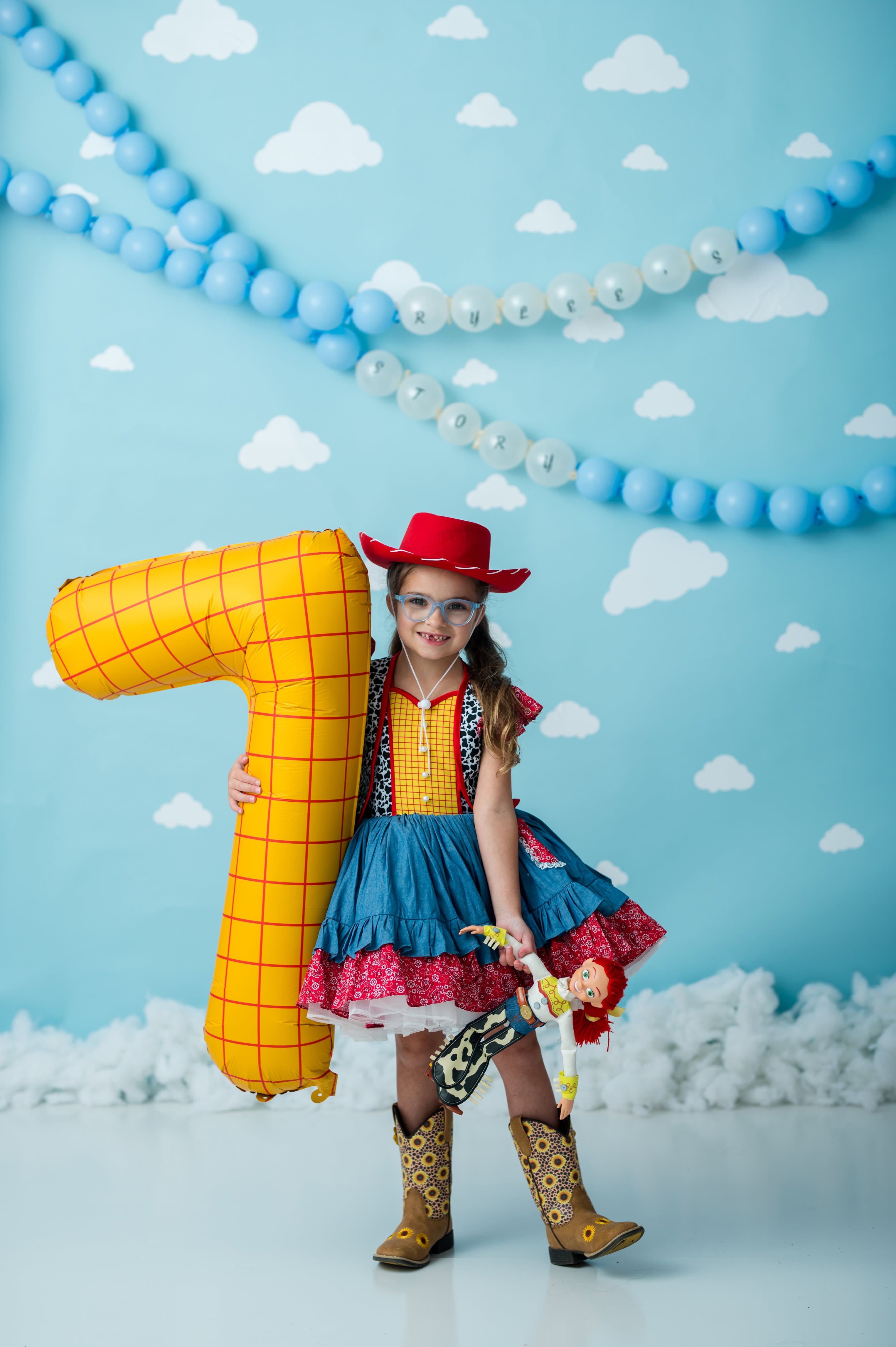 A young girl dressed as Woody from Toy Story stands in a decorated studio setting, holding a large yellow number 7 balloon. She is wearing a red cowboy hat, a blue and red dress, and brown cowboy boots against a light blue backdrop with white clouds and blue balloon garlands.