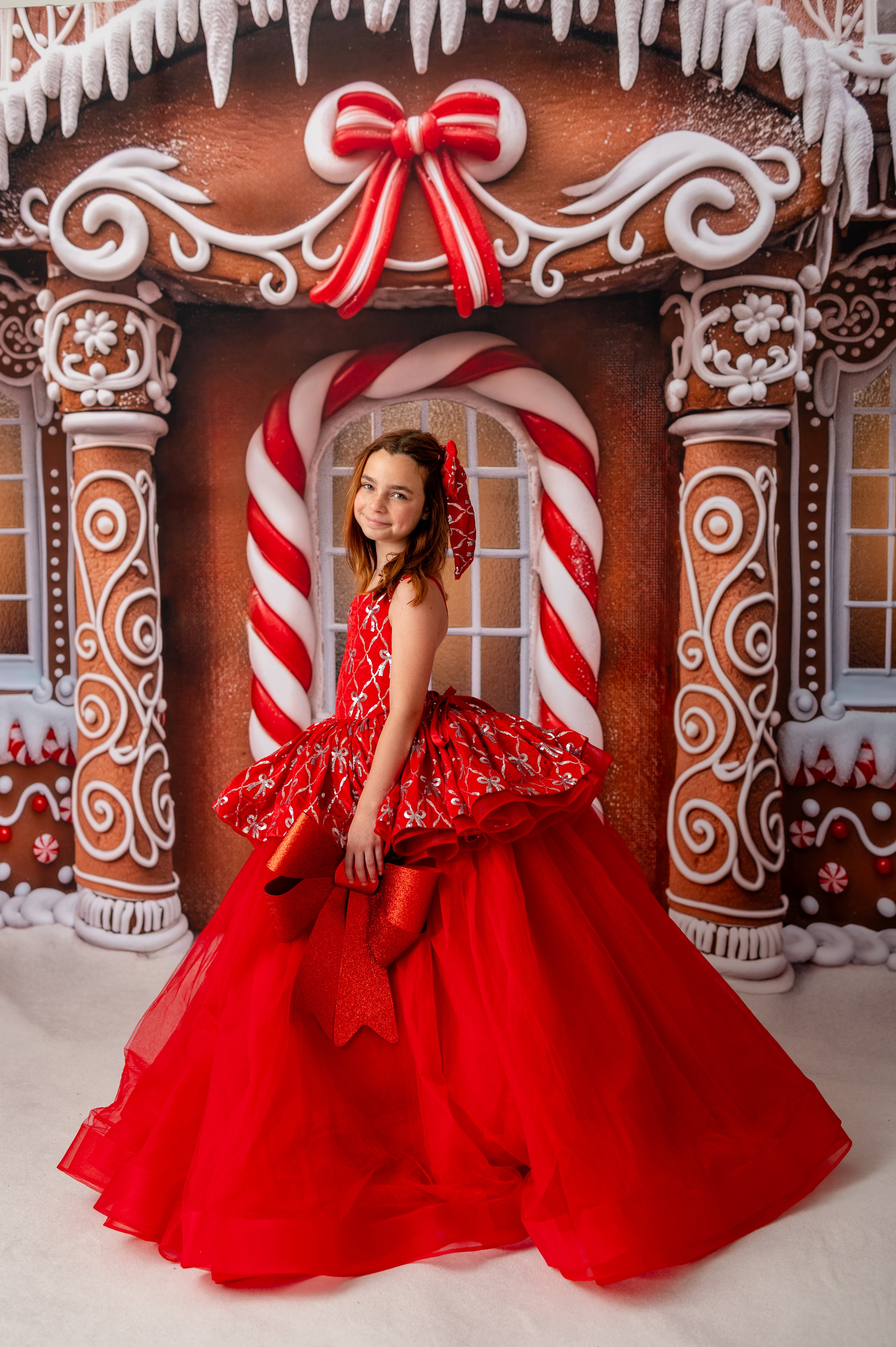 A young girl wearing a vibrant red velvet peplum gown with silver sequin bow detailing, standing in front of a whimsical gingerbread house backdrop.