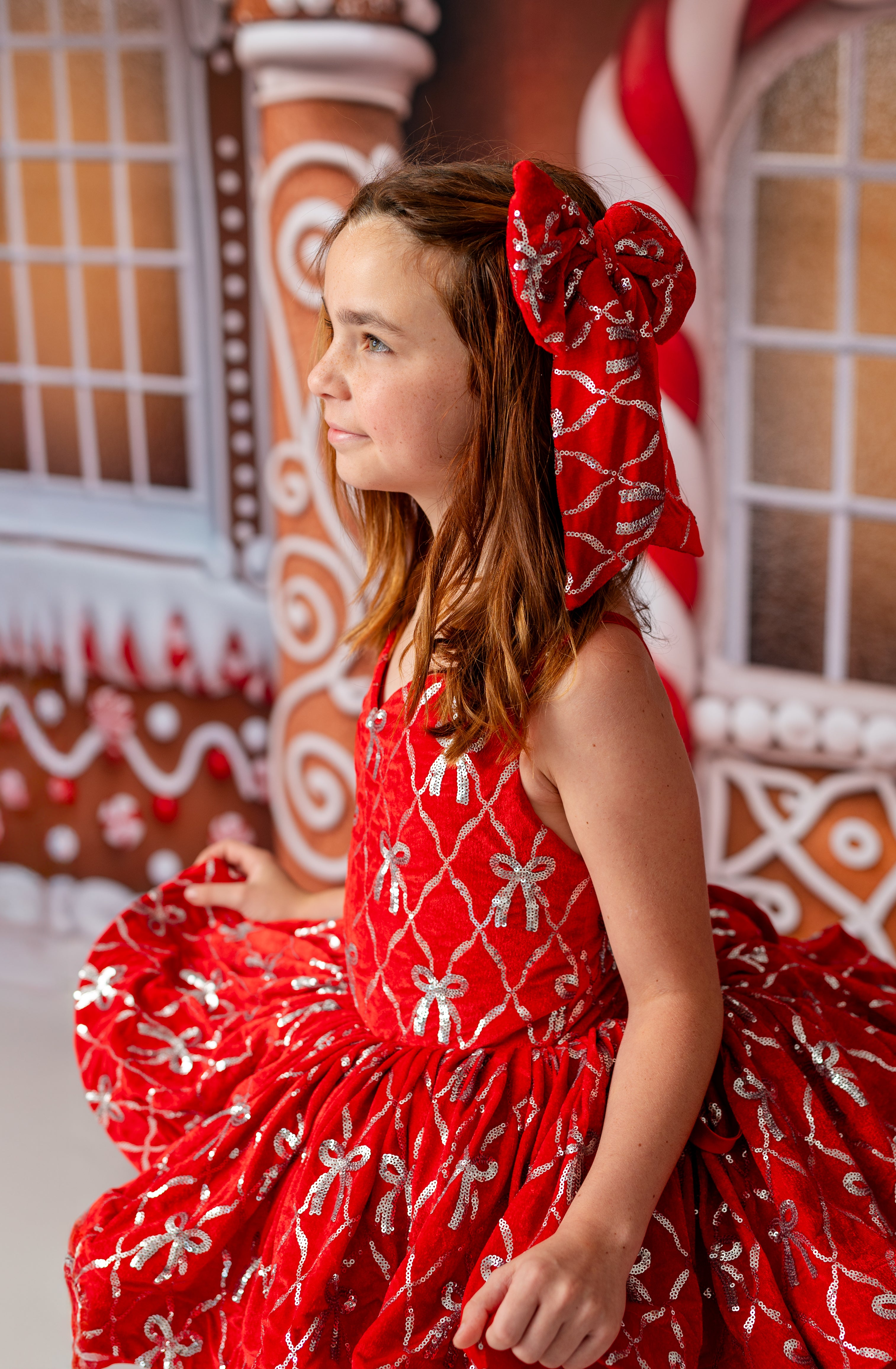 Young girl in a red dress with white patterns and a large bow in her hair, standing in front of a gingerbread house backdrop.