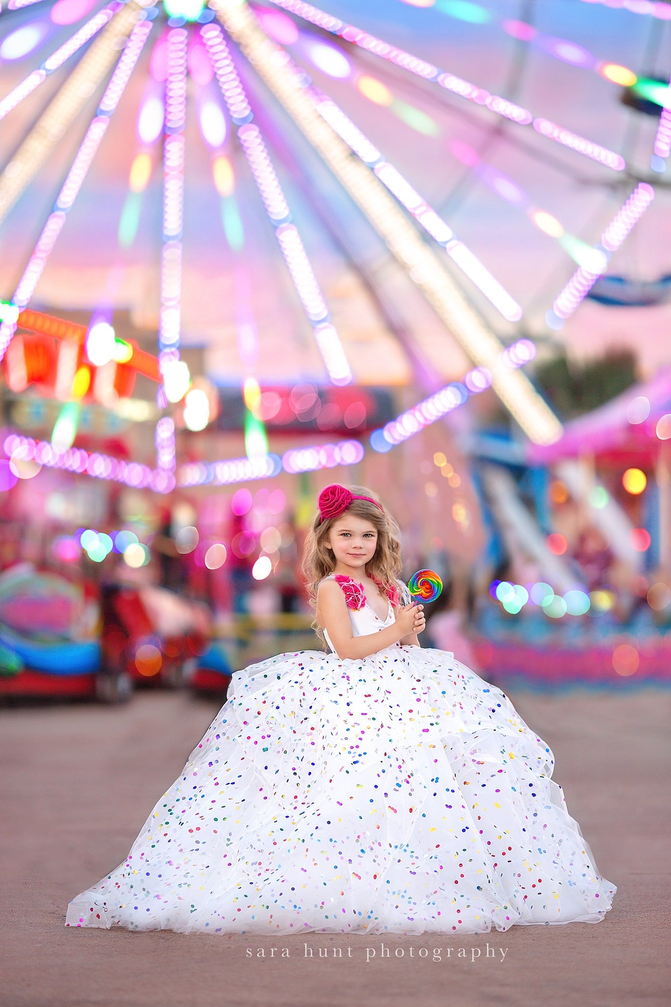 Girl in a confetti-dotted ball gown, standing at a fairground with vibrant lights in the background.