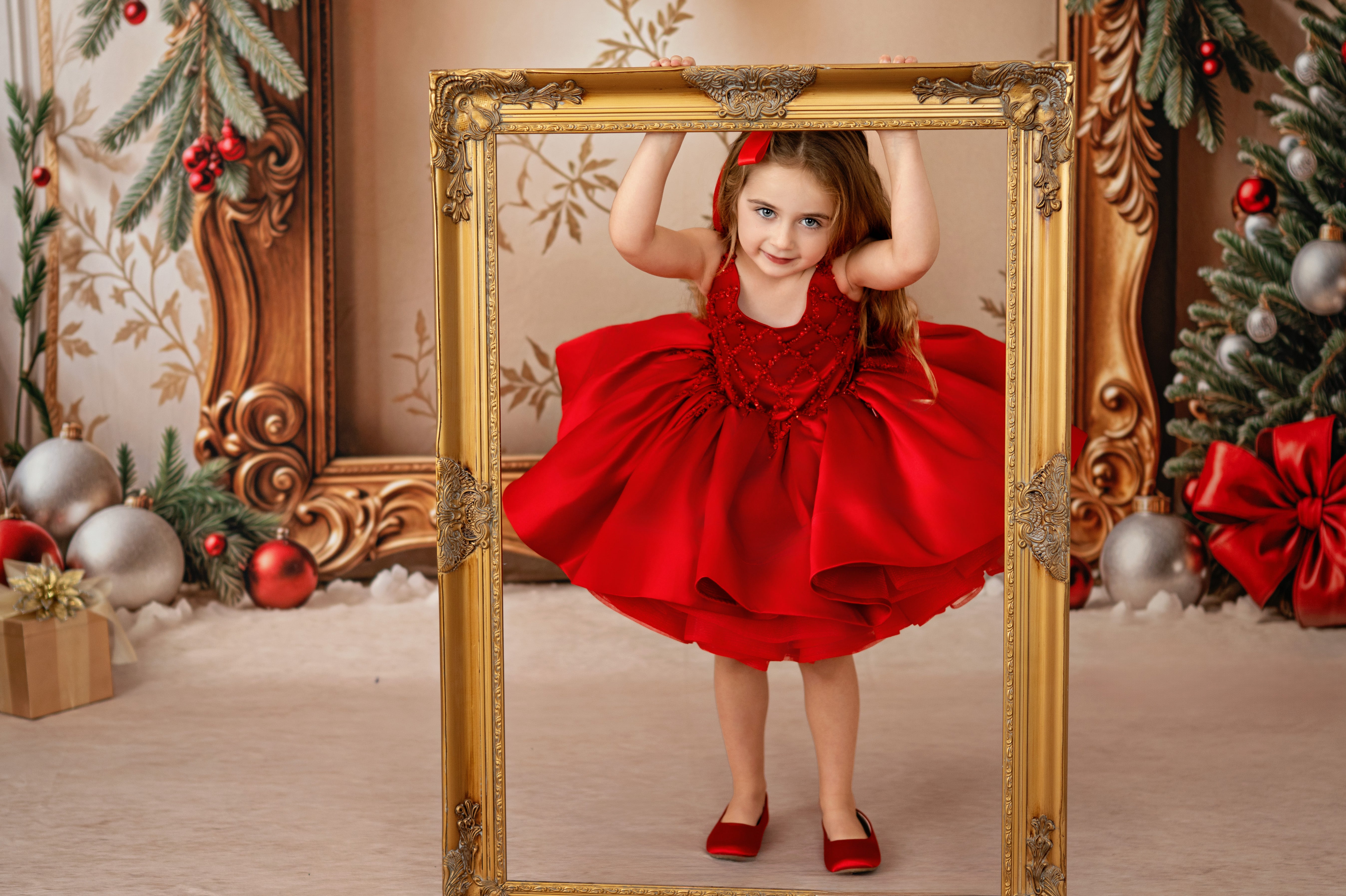 Young girl in a red dress standing in front of a gold ornate frame with Christmas decorations in the background.