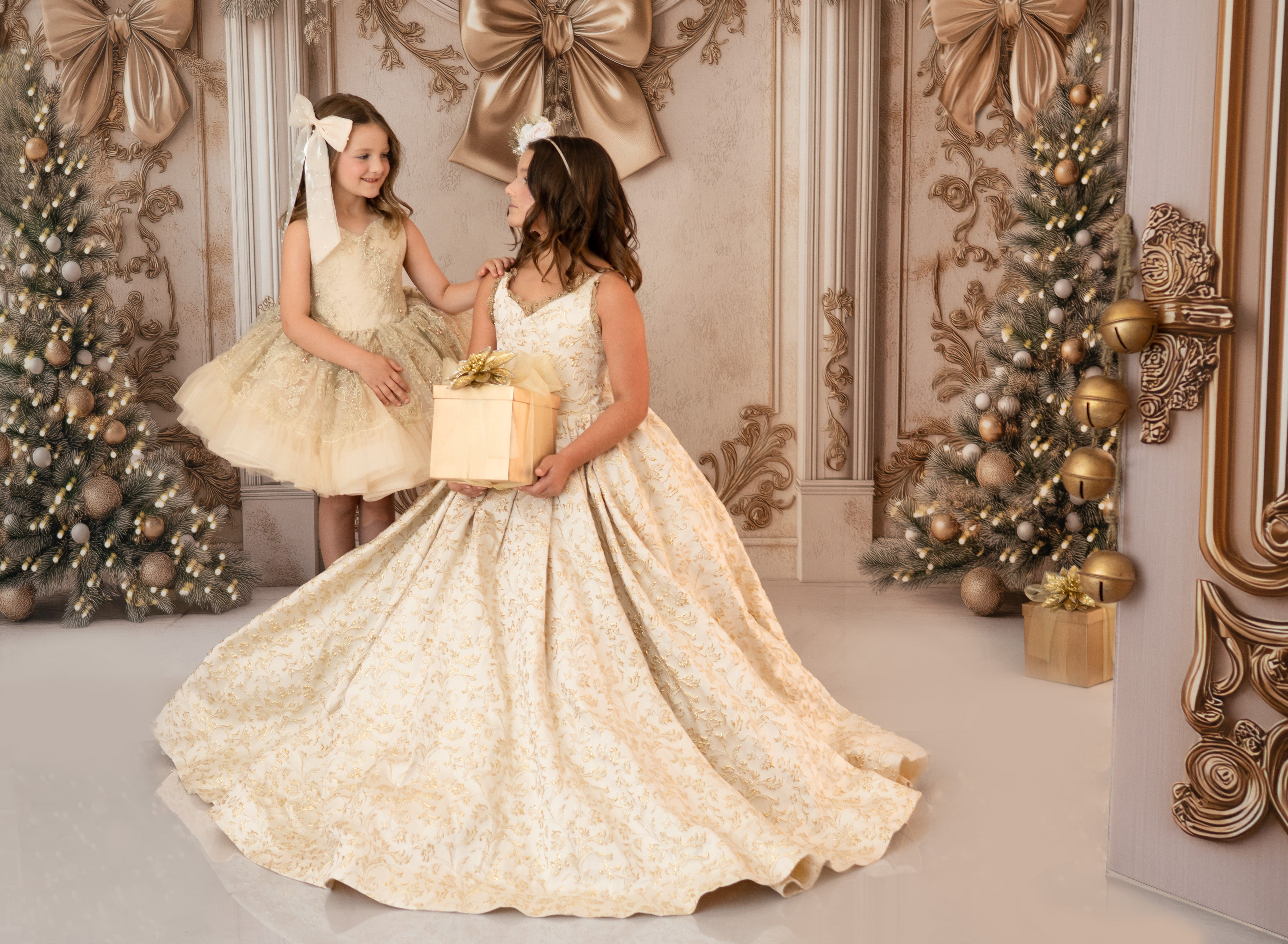 Two young girls in elegant dresses holding a gift box in a decorated room with Christmas trees.