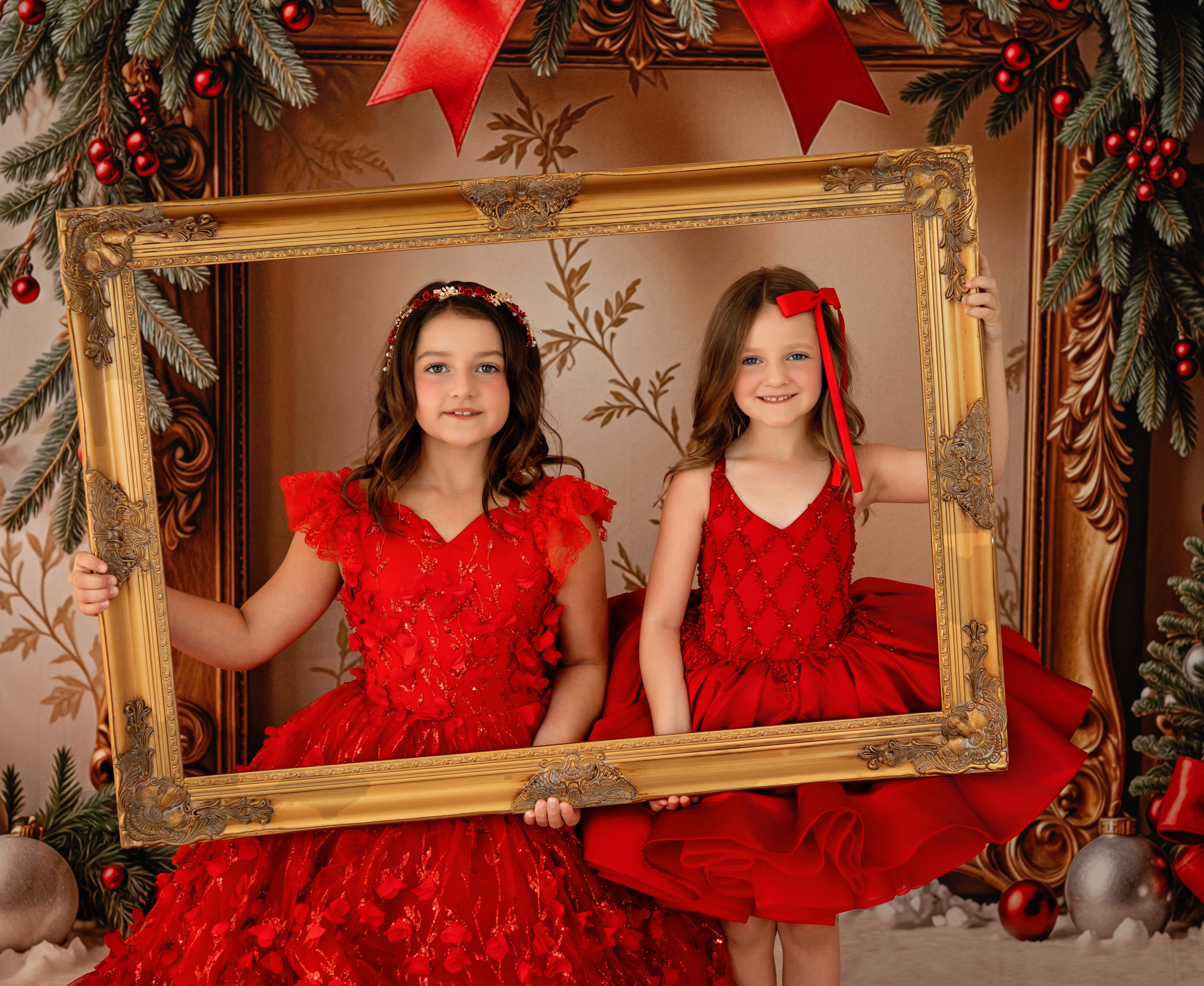 Two girls in red dresses holding a gold-framed mirror decorated with Christmas elements.
