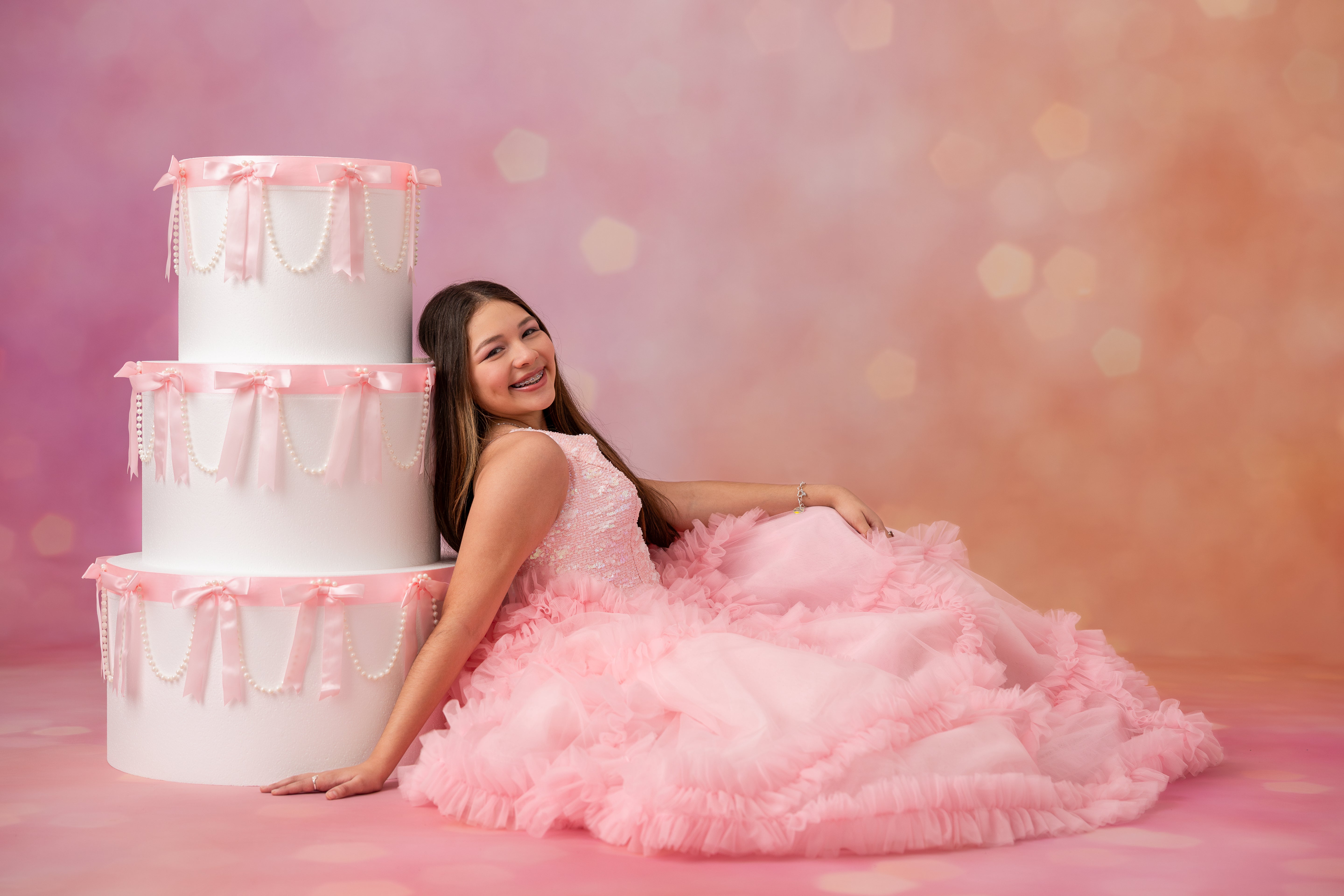 Woman in a pink dress sitting next to a large white cake with pink ribbons against a pink background