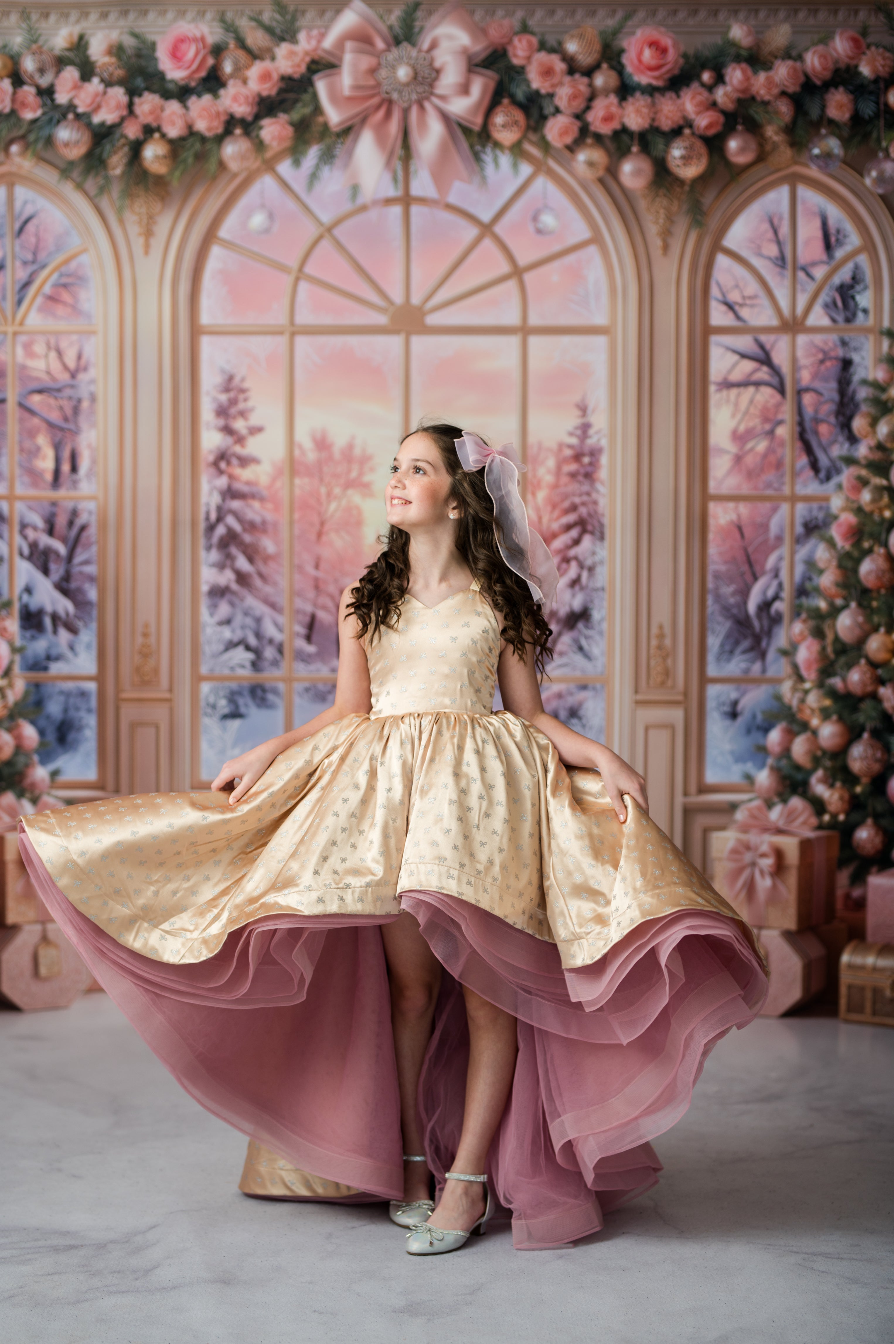 A young girl stands in a festive holiday-themed studio setting wearing a formal ballgown. The dress features a champagne-gold satin bodice and skirt with a layered dusty pink tulle overskirt and matching veil.