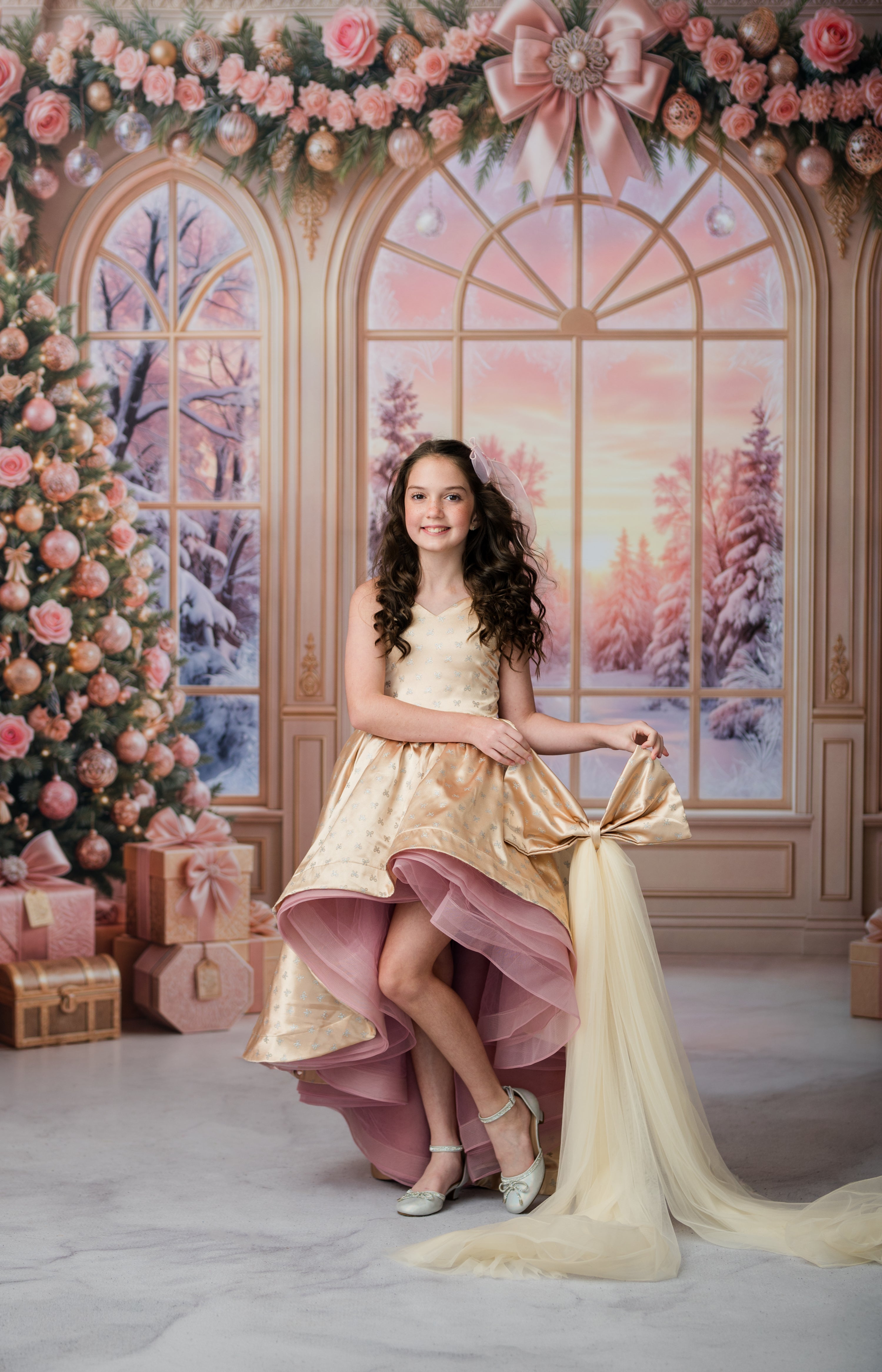 A young girl with long curly brown hair stands in a festive holiday studio setting, wearing a champagne-gold and dusty pink high-low dress with a large bow at the back. The background features a Christmas tree, large arched windows with a winter sunset scene, and pink rose garlands.