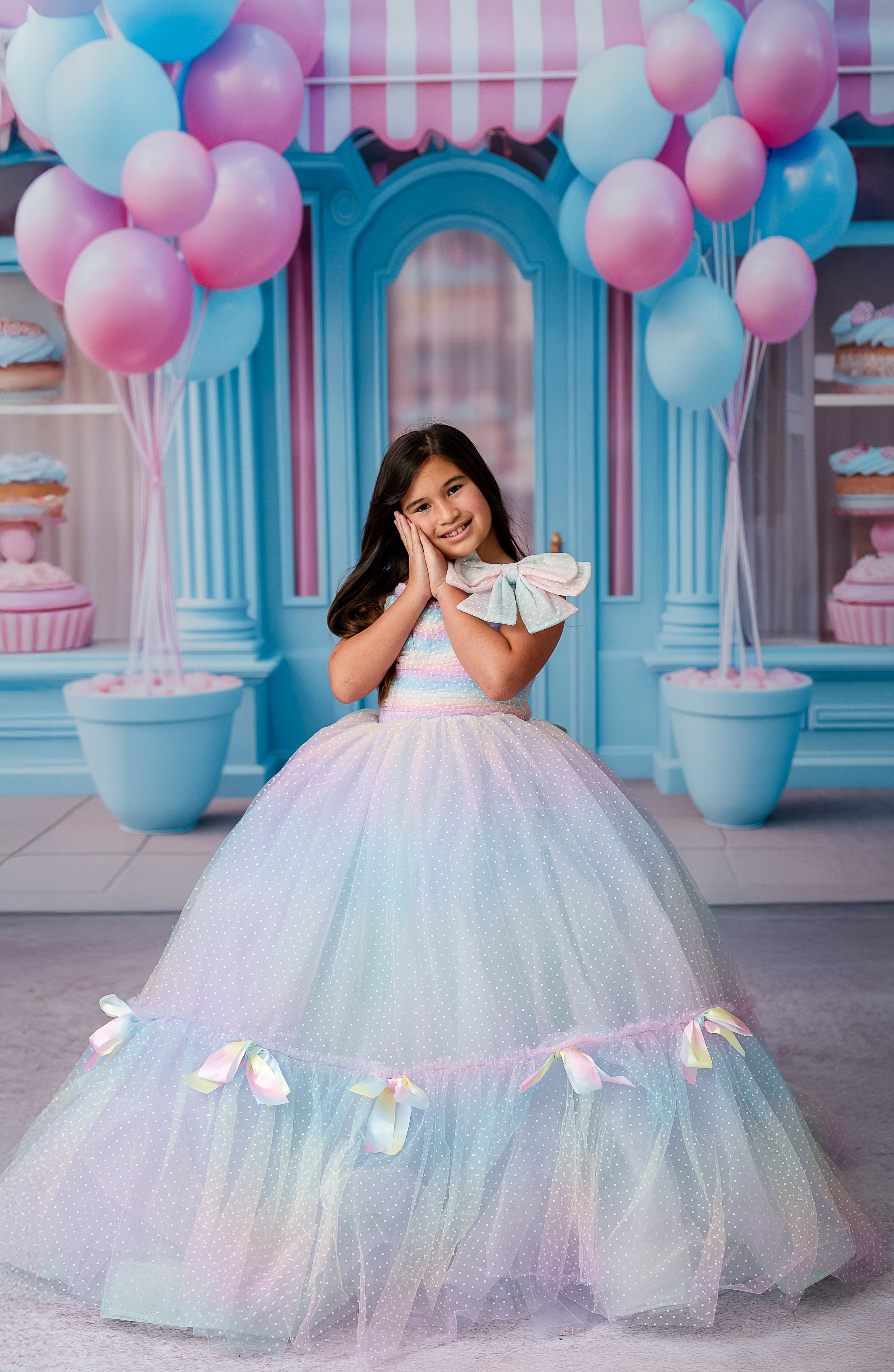 Child wearing the Colorful Bliss Gown, showcasing a floor-length rainbow swiss dot fabric with soft pastel tones and satin bows along the hemline.