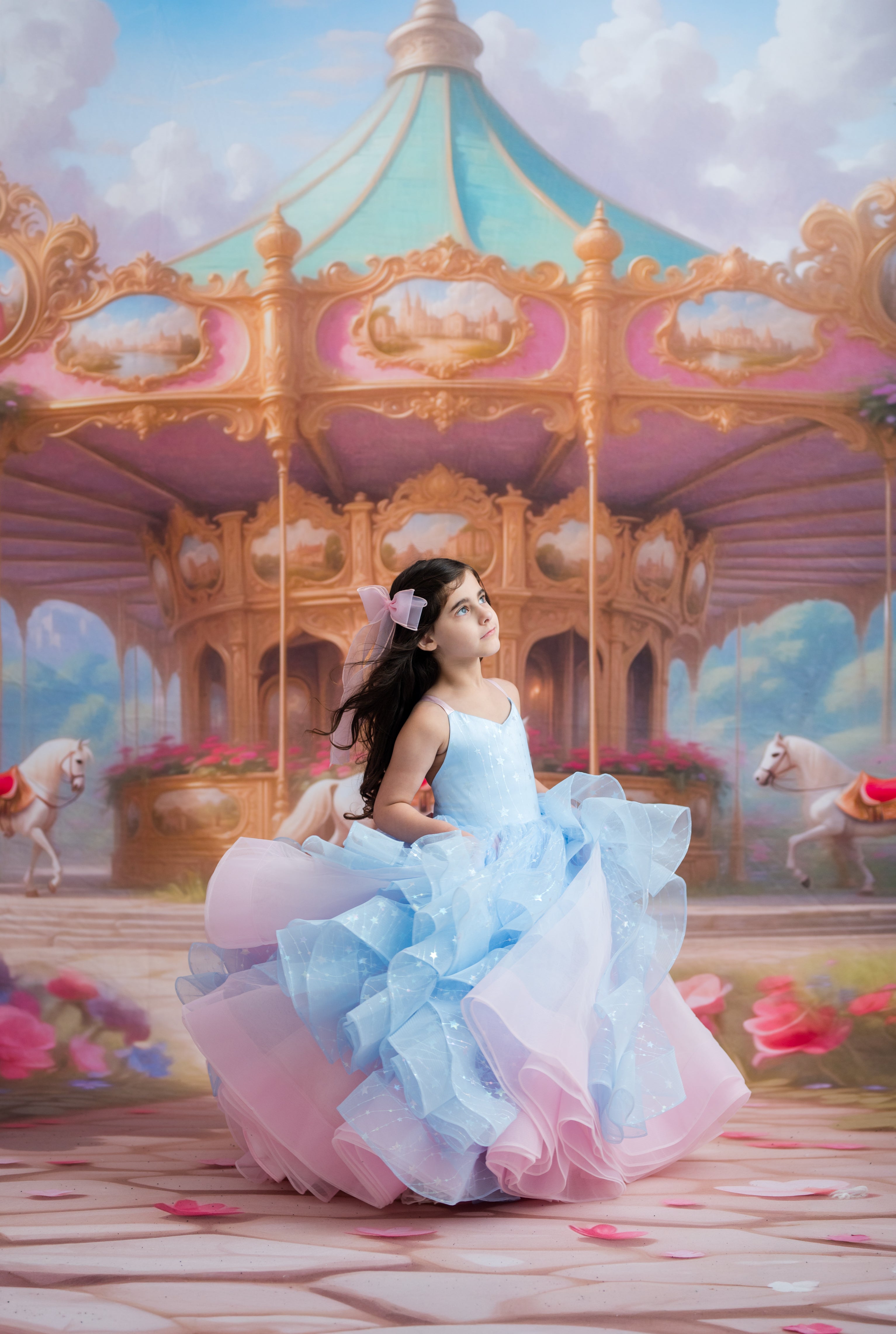 A young girl in a voluminous light blue and pink ballgown sits on a carousel in front of a whimsical pastel carousel scene.