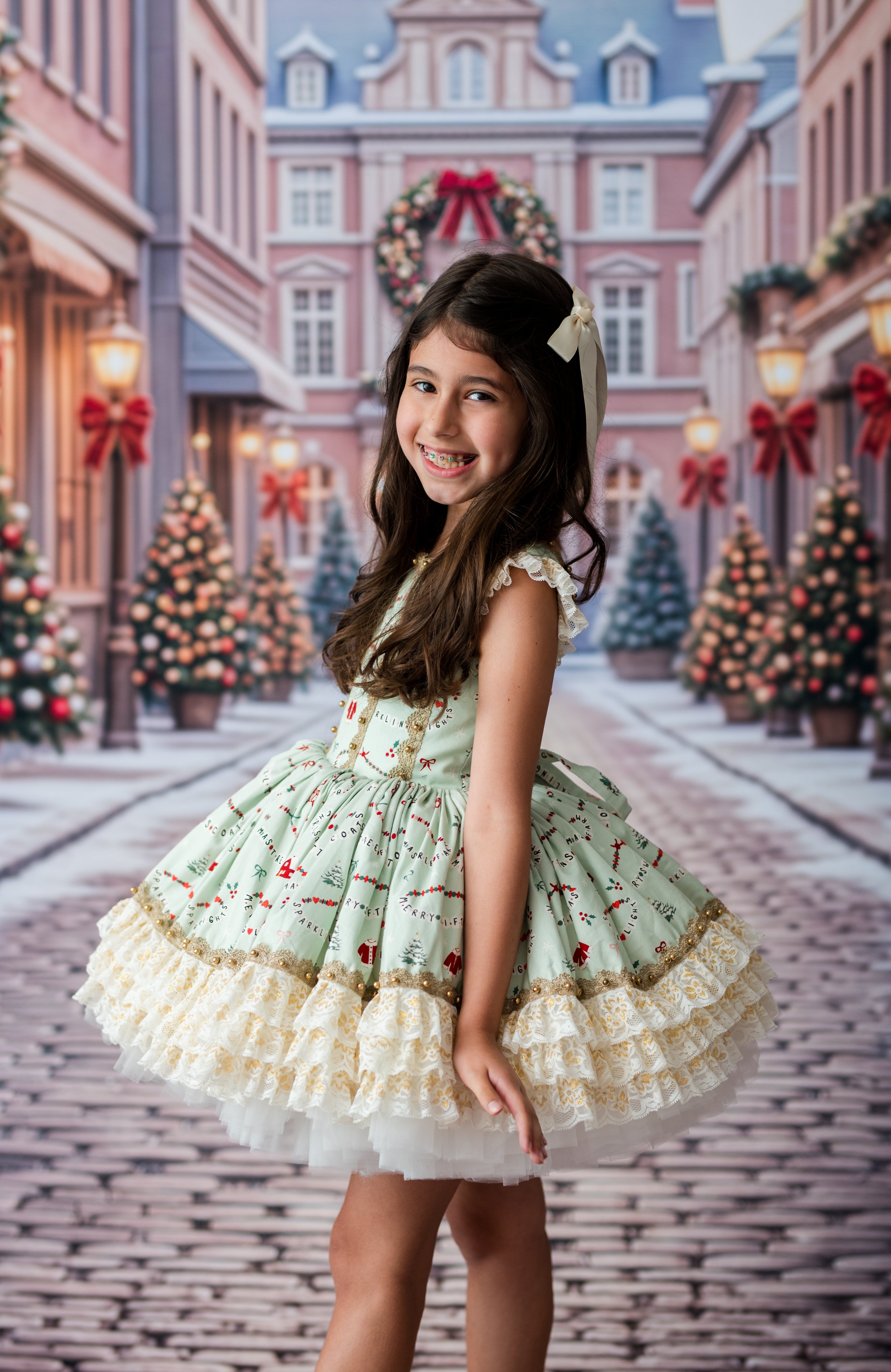 A young girl with long brown hair stands in a festive, snowy town street wearing a light green holiday dress with cream ruffles and a white bow headband.