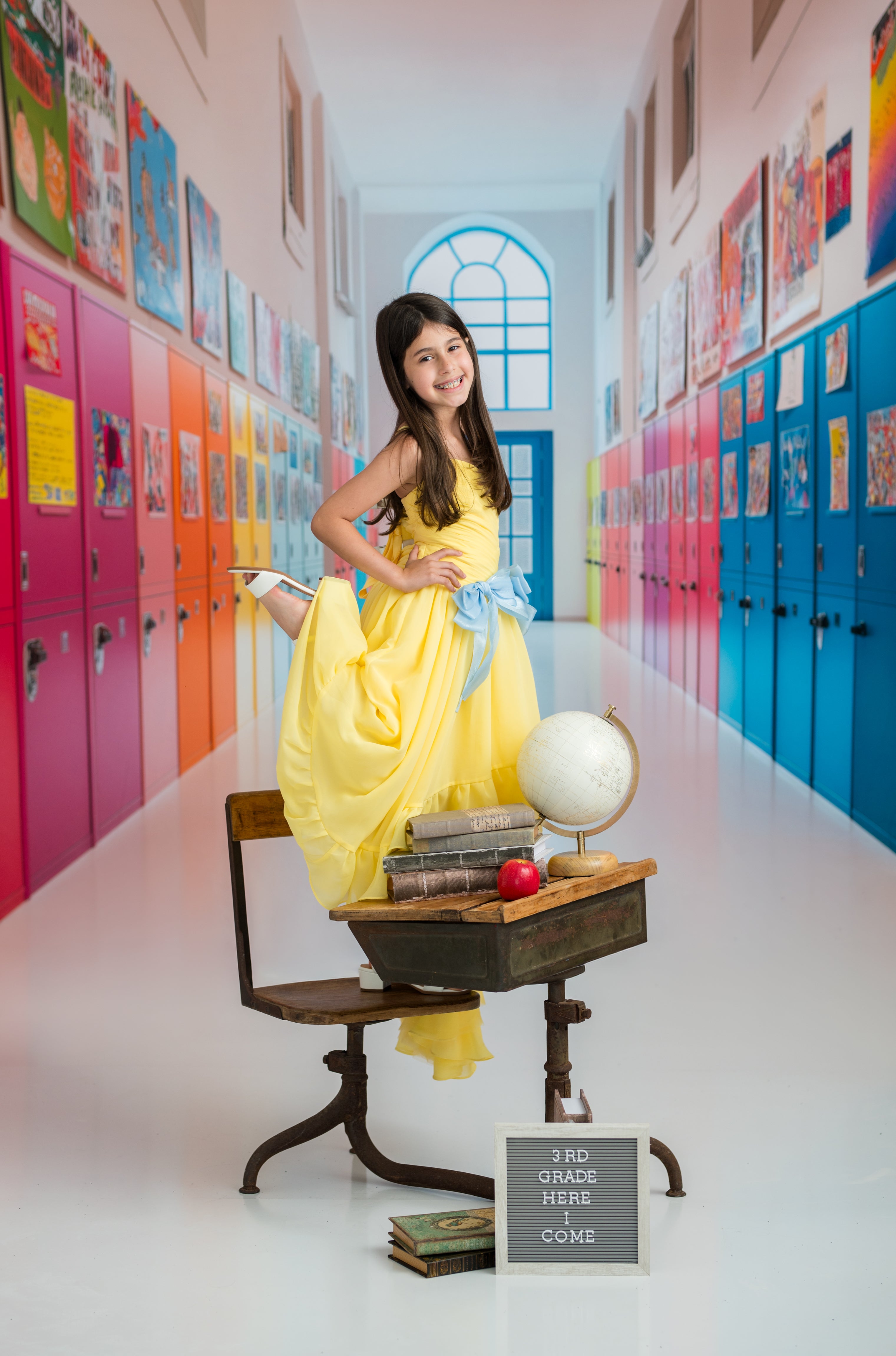 Young girl in a yellow dress standing in a colorful school hallway with lockers.