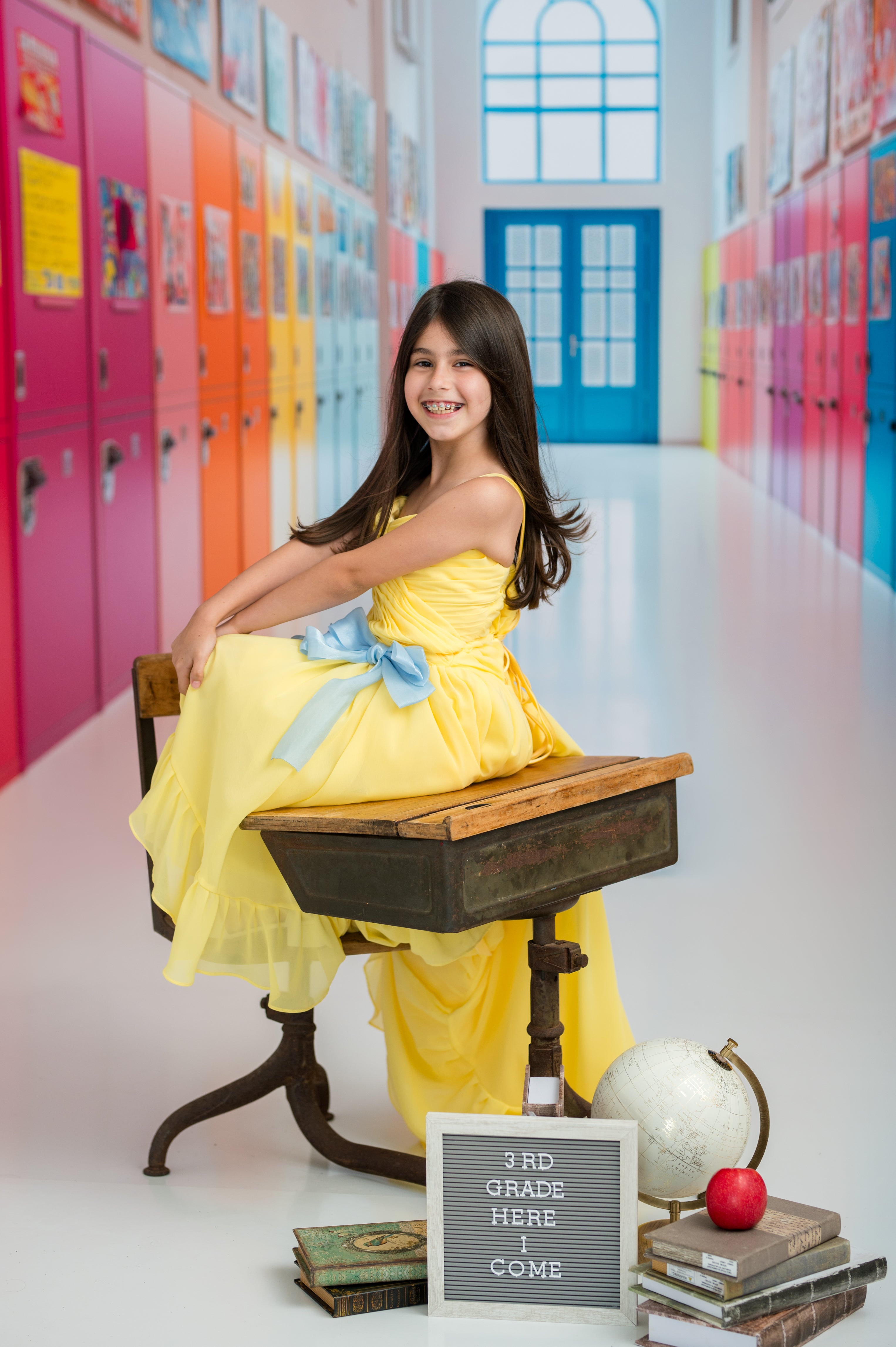 Young girl in a yellow dress sitting on a desk in a colorful school hallway.