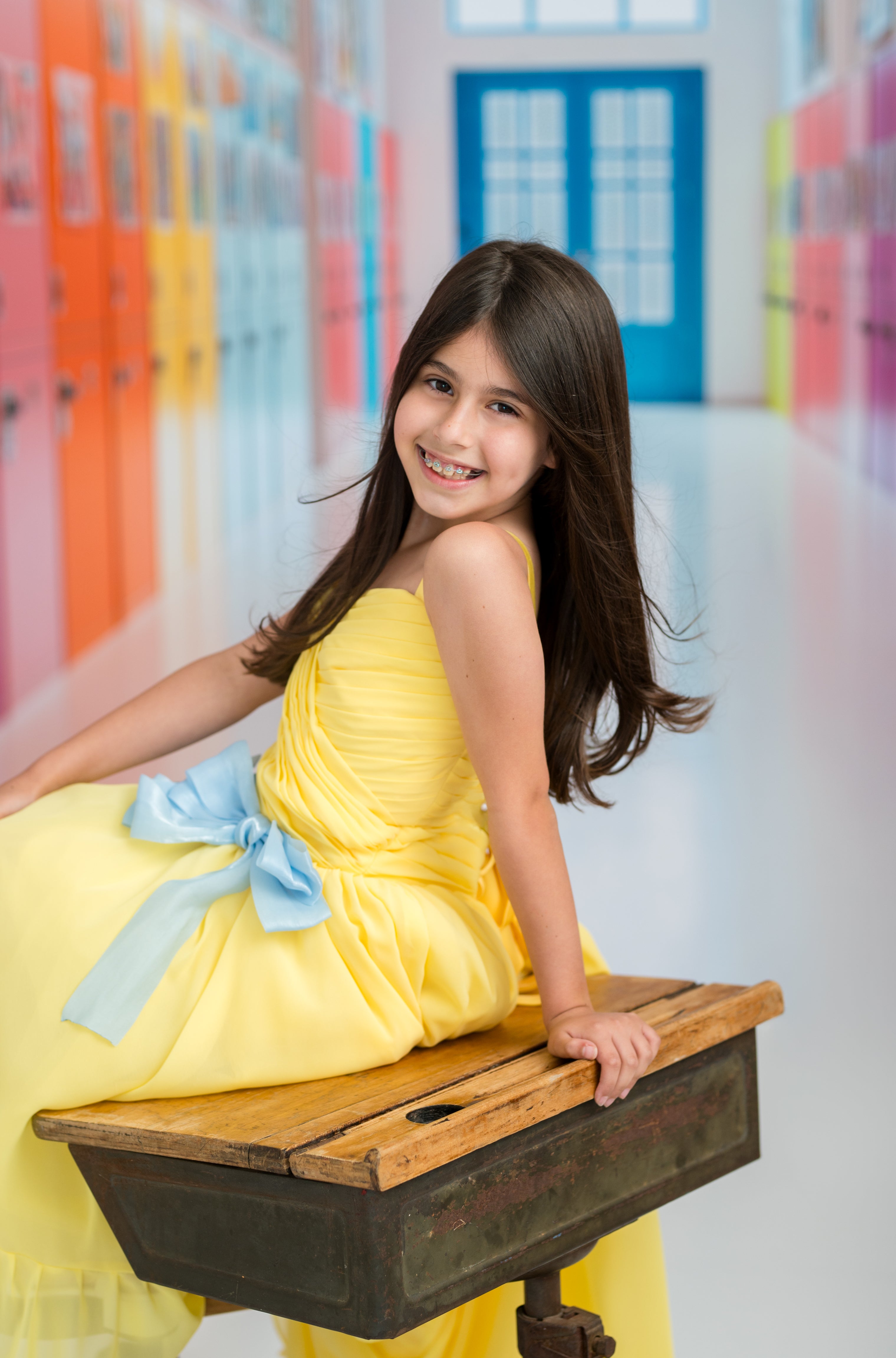 Young girl in a yellow dress with a blue bow sitting on an old school desk in a colorful hallway.
