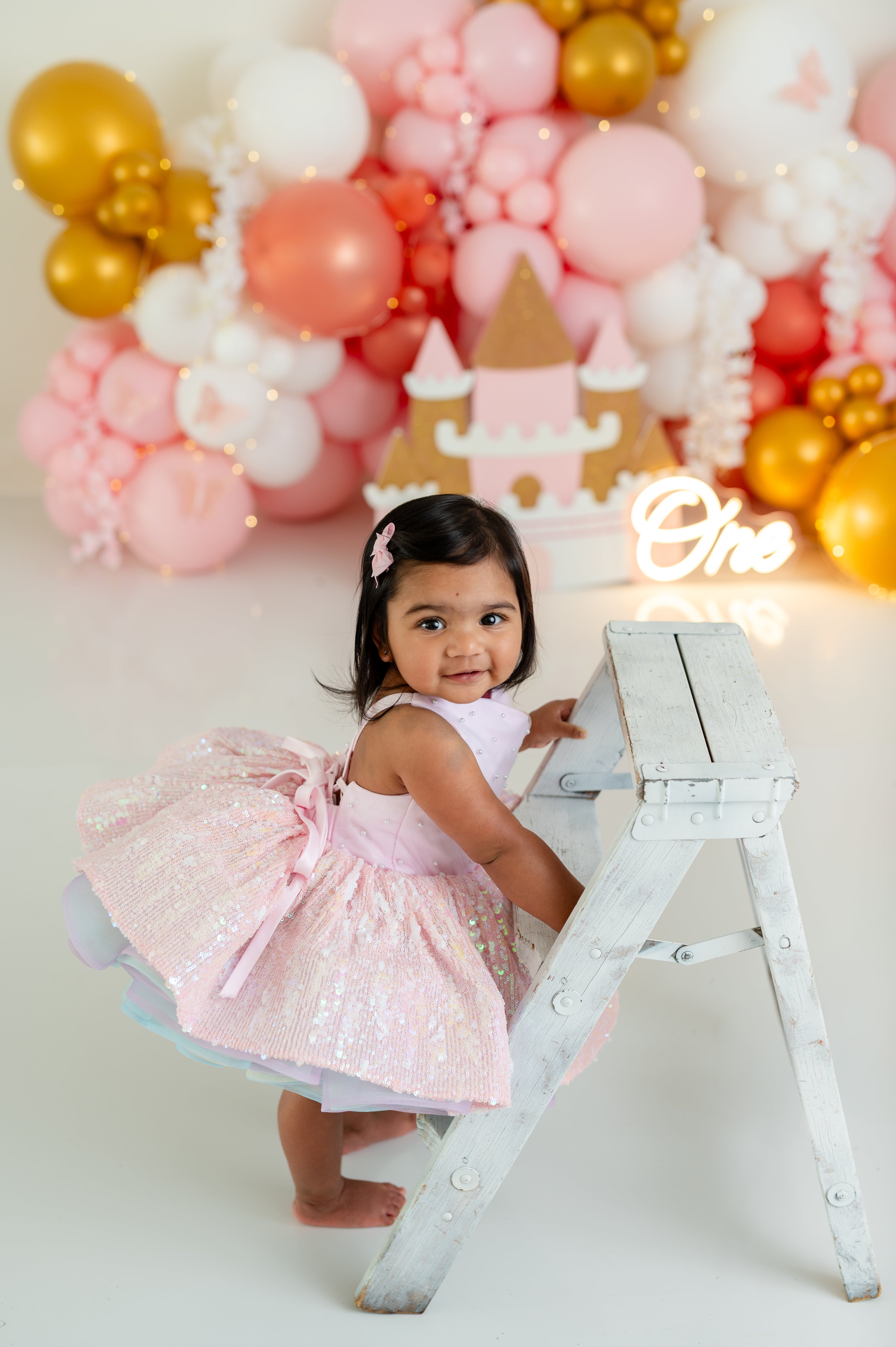 Child in a pink dress standing on a small white ladder with a colorful balloon arch and castle decoration in the background.