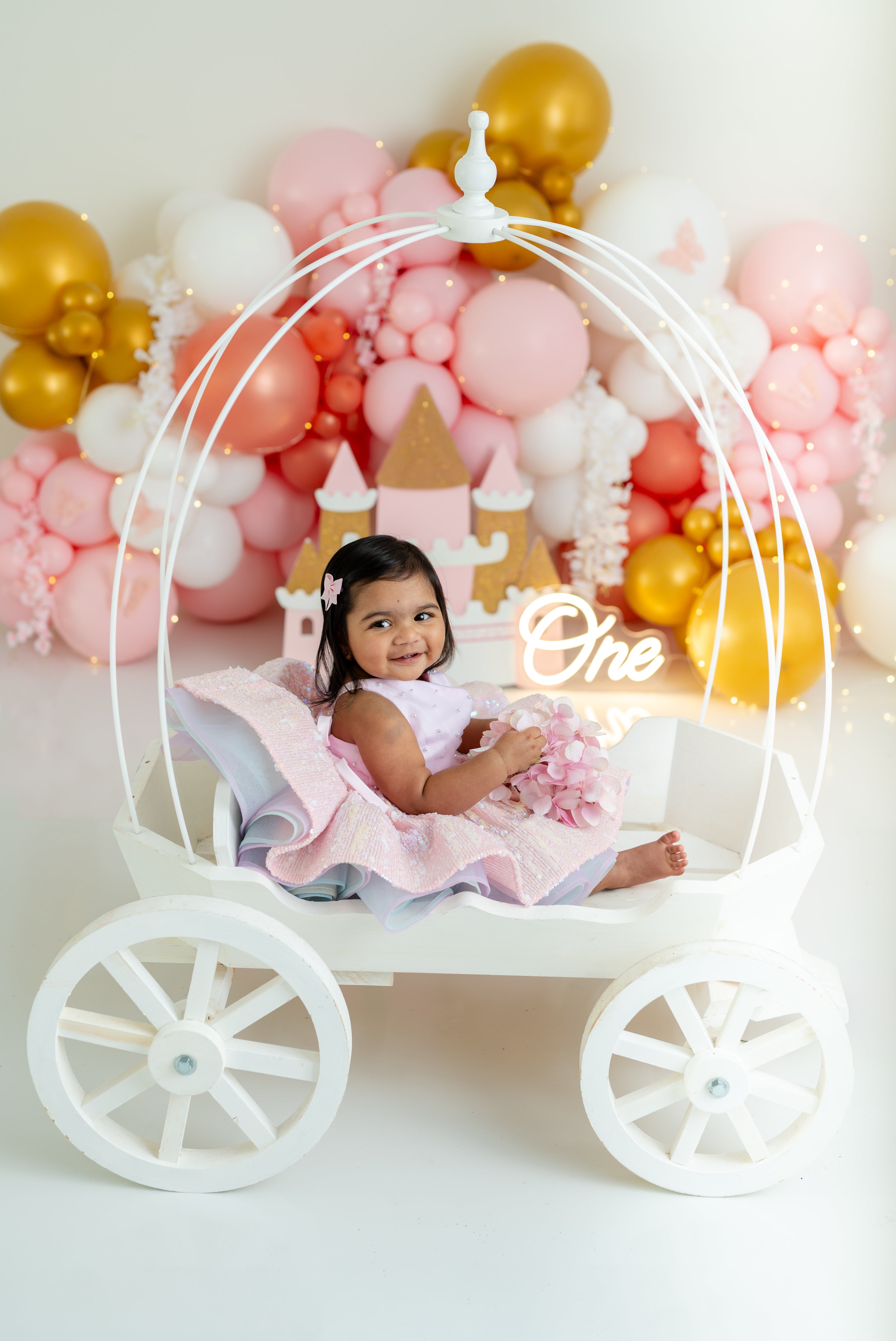 Child in a decorated carriage with balloons and 'One' sign, celebrating a birthday.