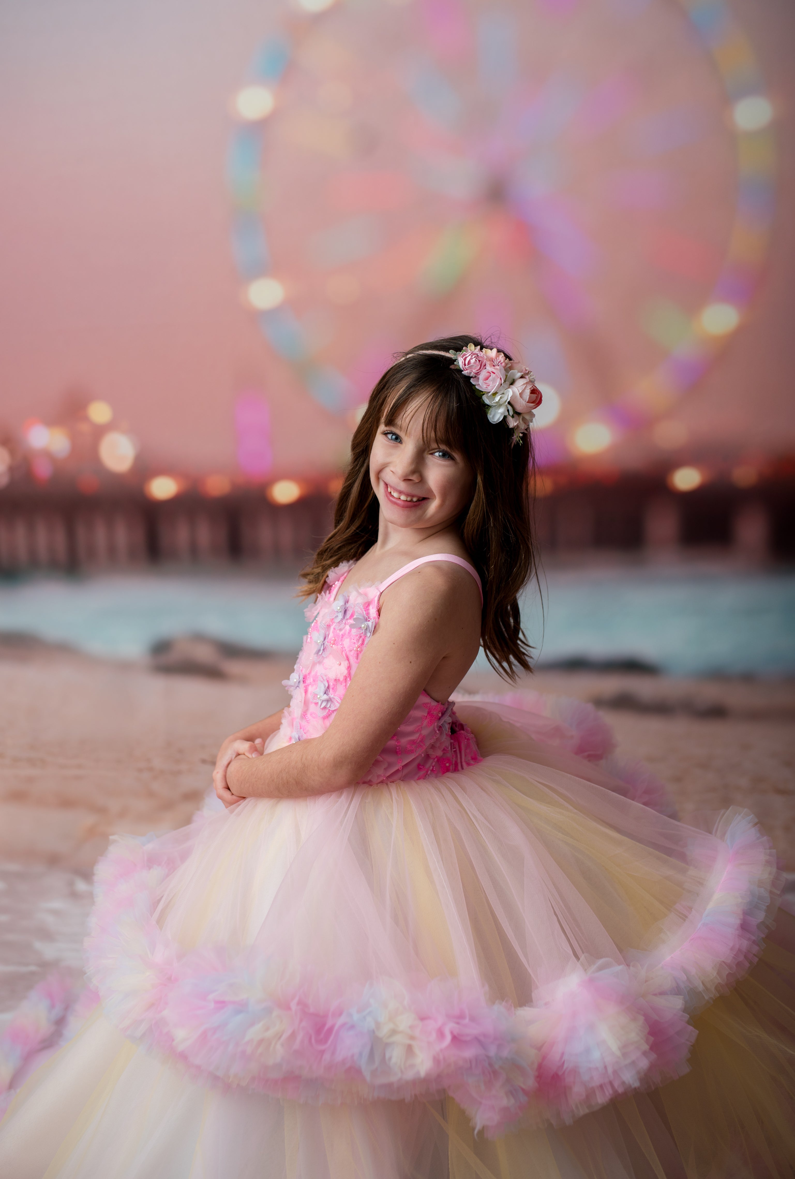 Young girl in a pink and yellow dress with flowers, standing in front of a Ferris wheel at sunset.