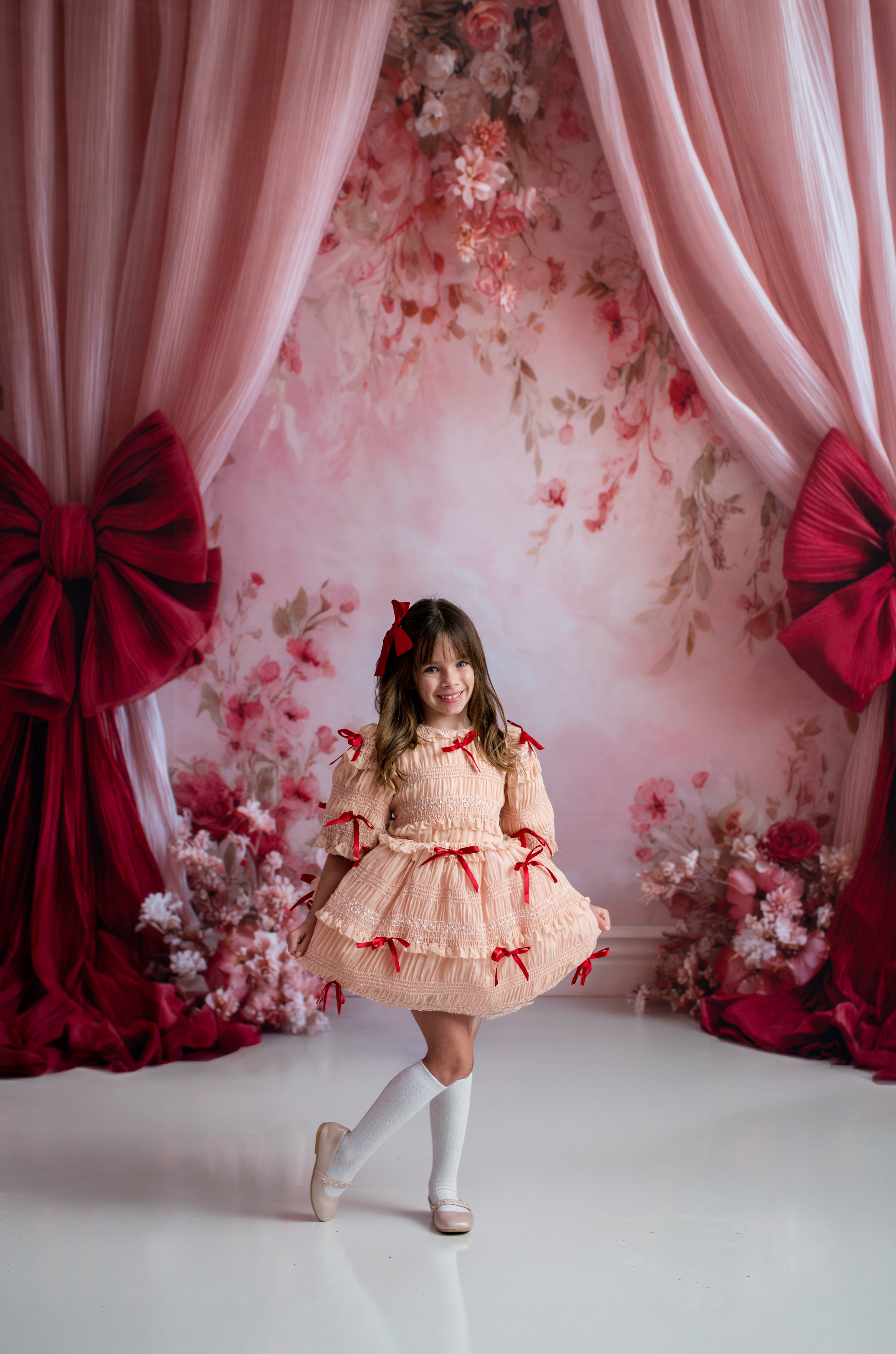 Young girl in a peach dress with red ribbons standing in front of a floral and curtain backdrop.