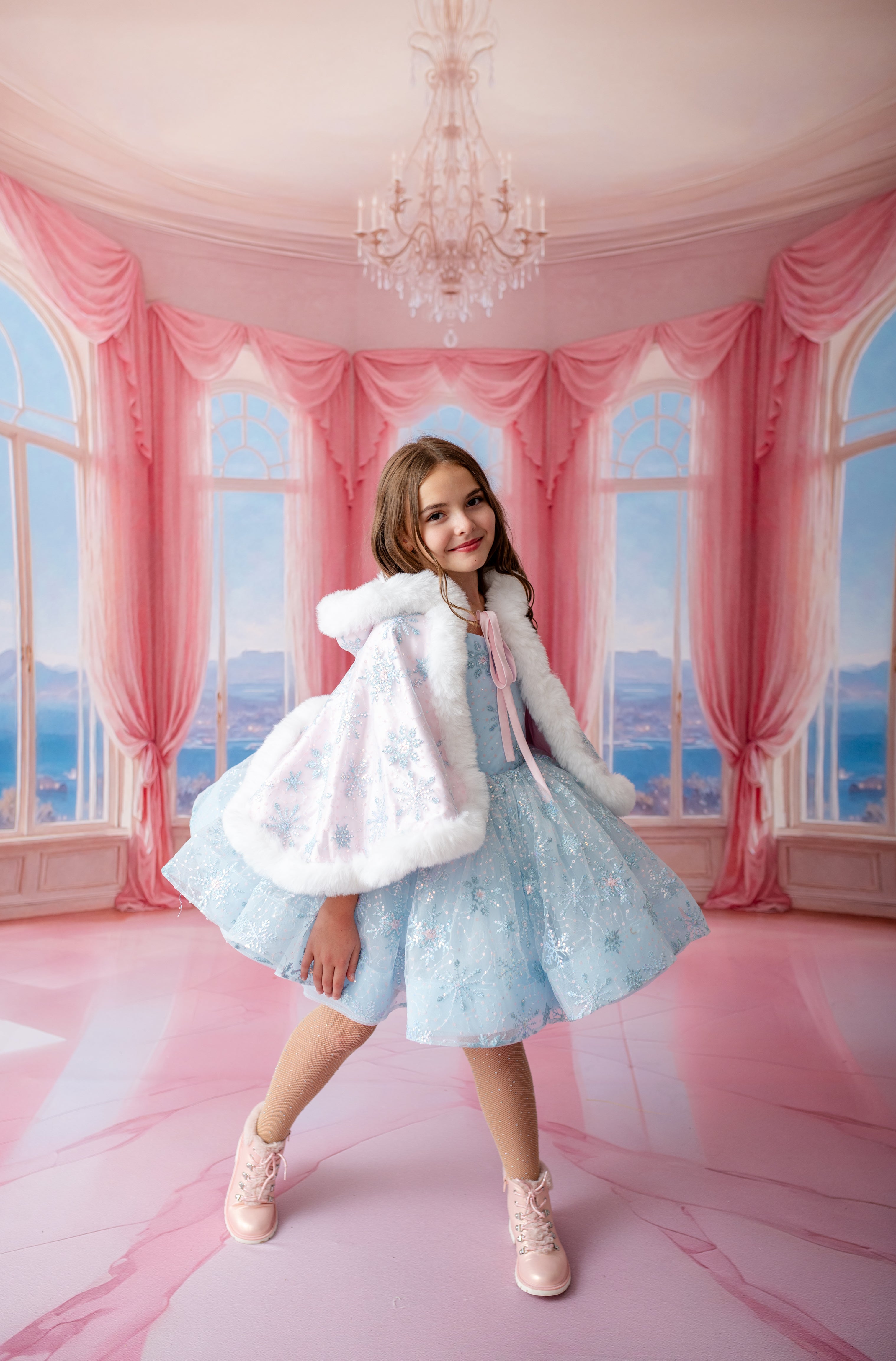 Young girl in a light blue dress with white fur trim standing in a room with pink curtains and a chandelier.