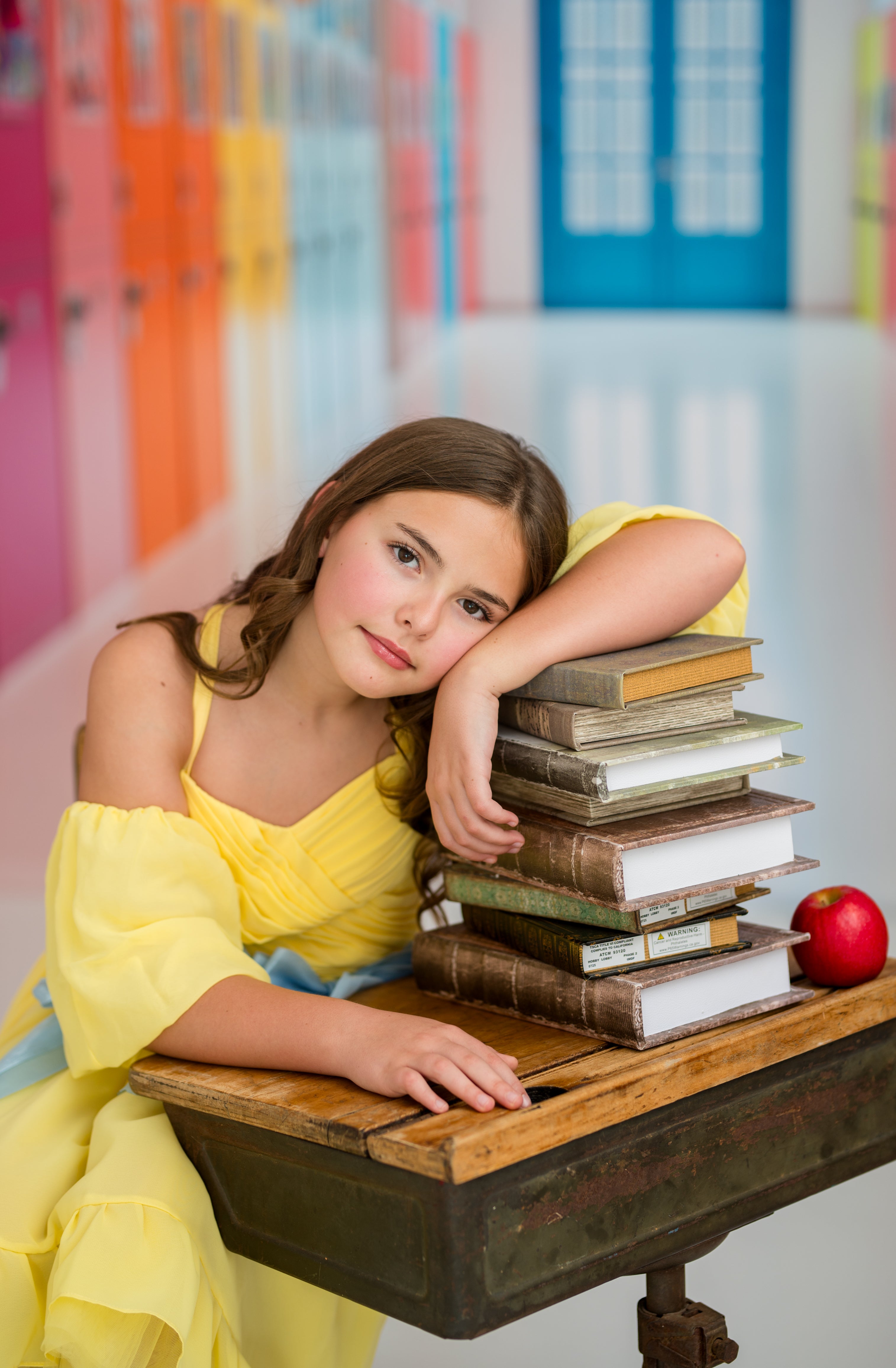 Young girl in a yellow dress sitting at a desk with books and an apple, surrounded by colorful bookshelves.