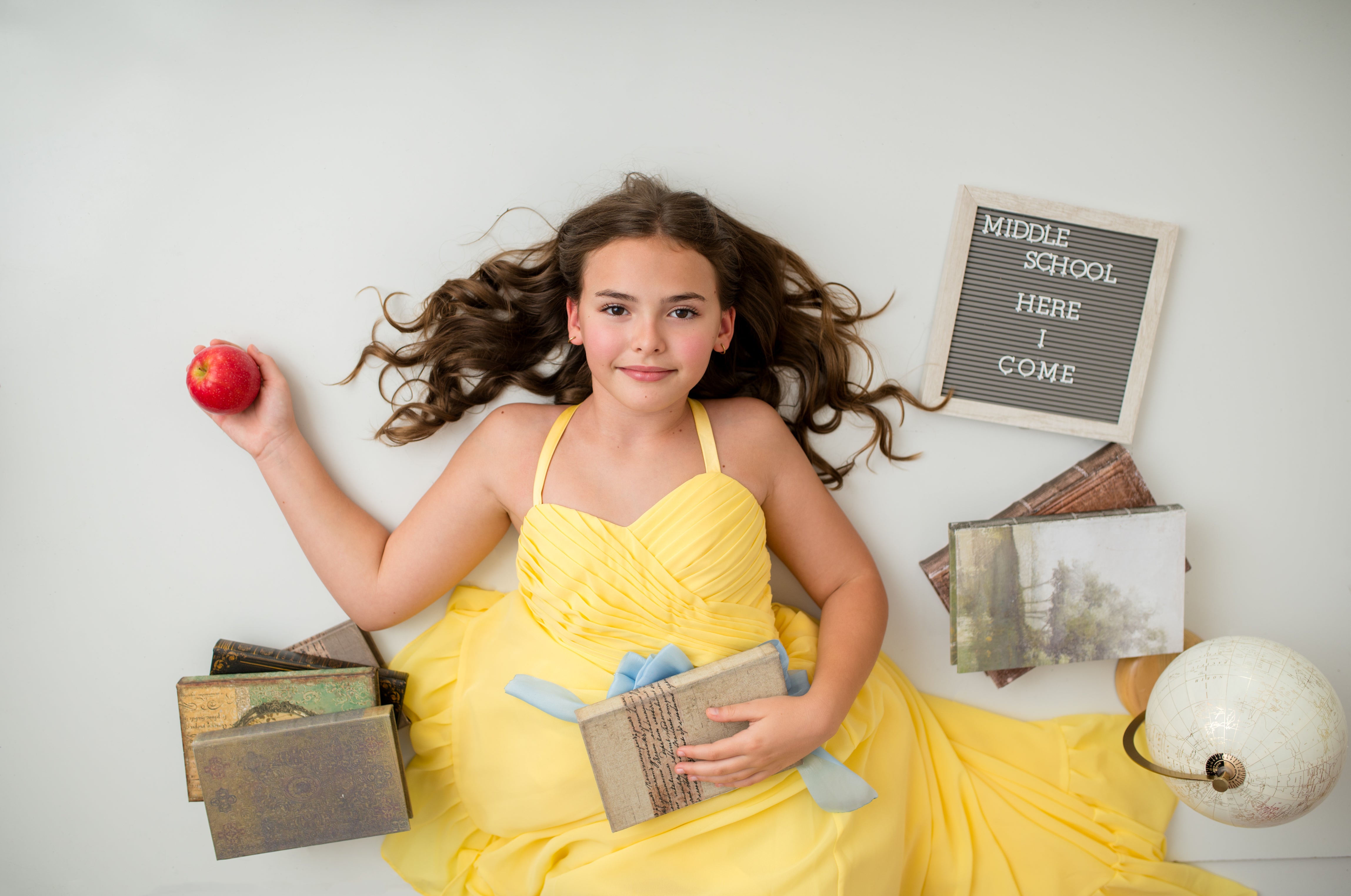 Young girl in a yellow dress holding books and an apple, with a 'Middle School Here I Come' sign in the background.