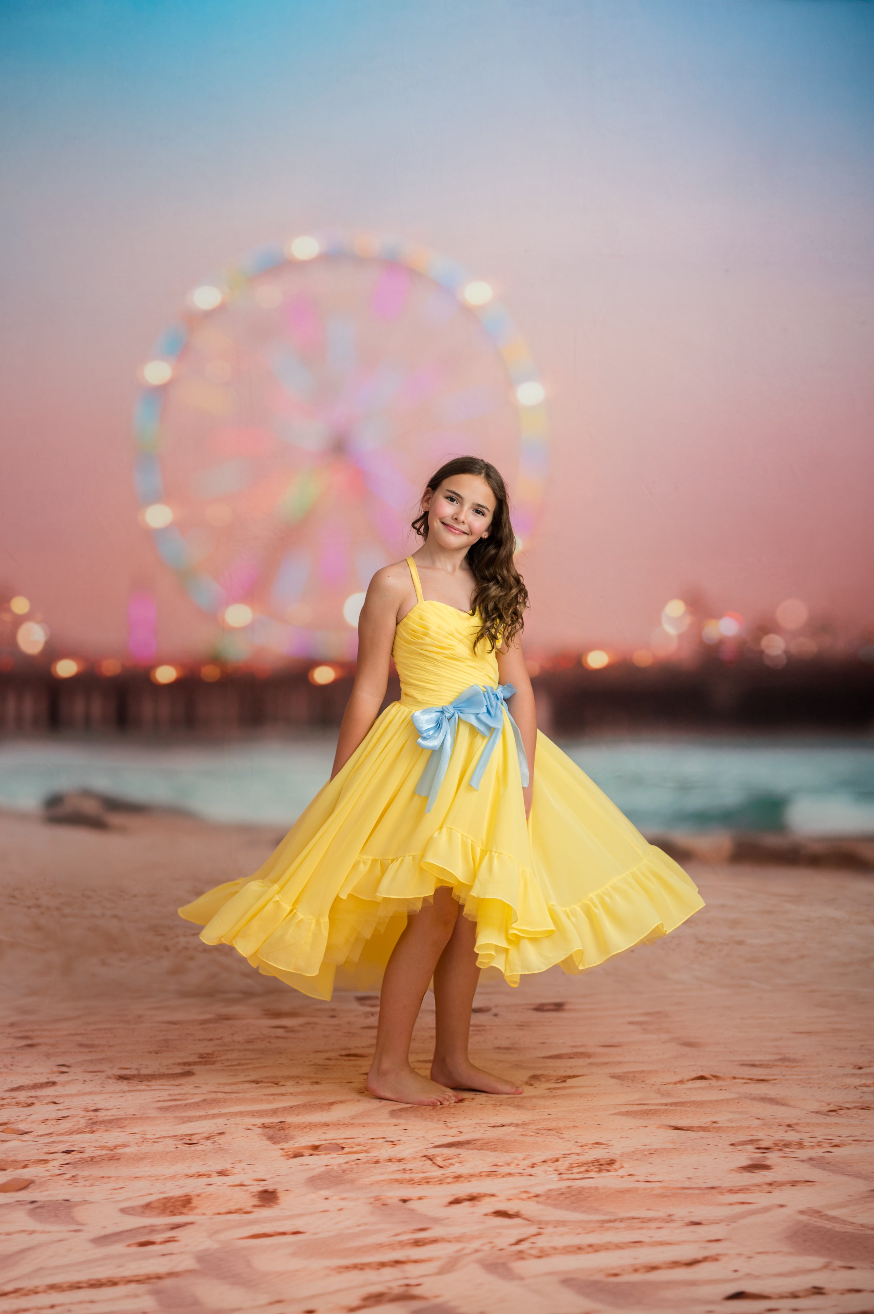 Young girl in a yellow dress with a blue bow standing on a beach with a Ferris wheel in the background.