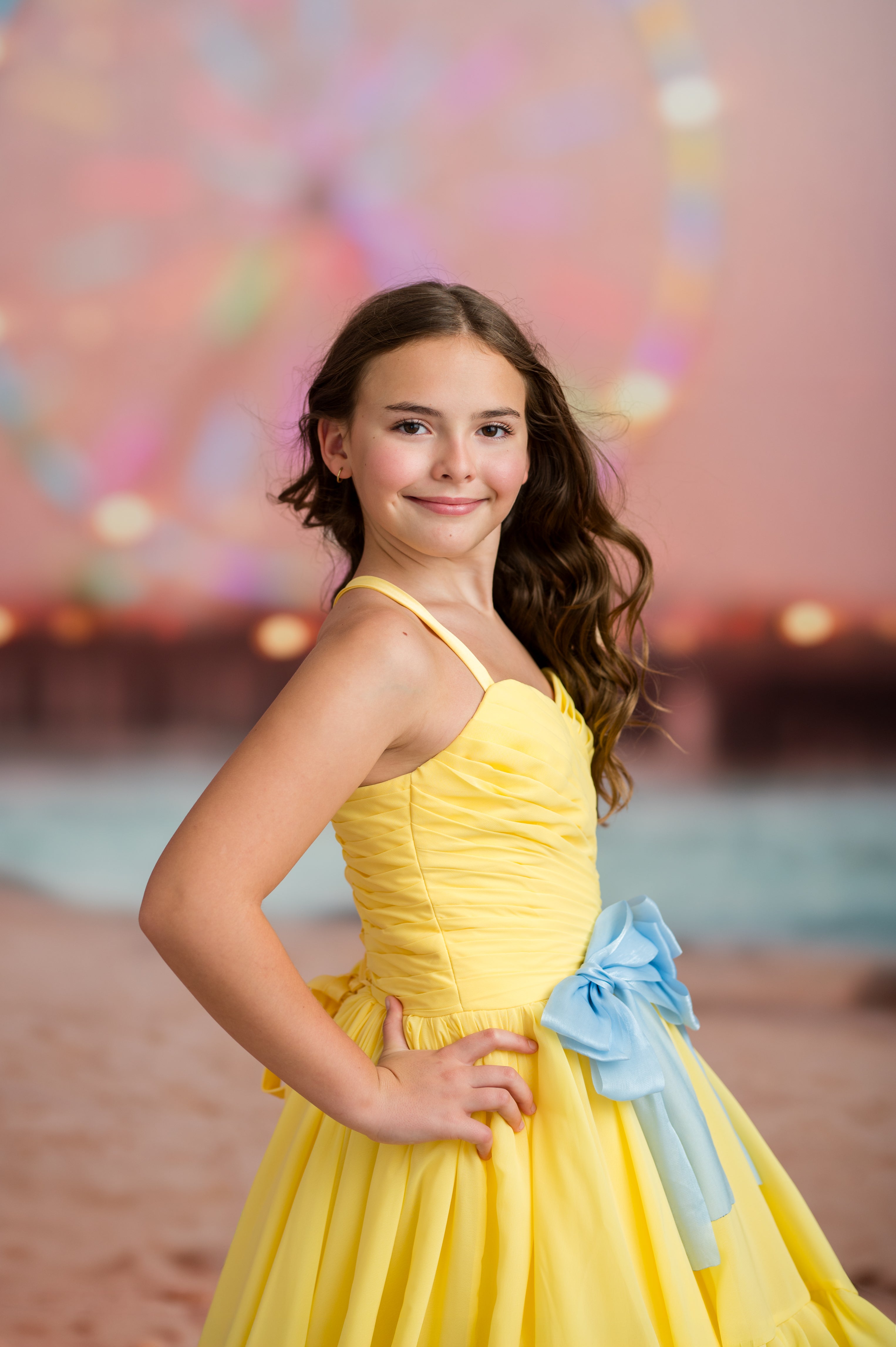 Young girl in a yellow dress with a blue bow standing on a beach.