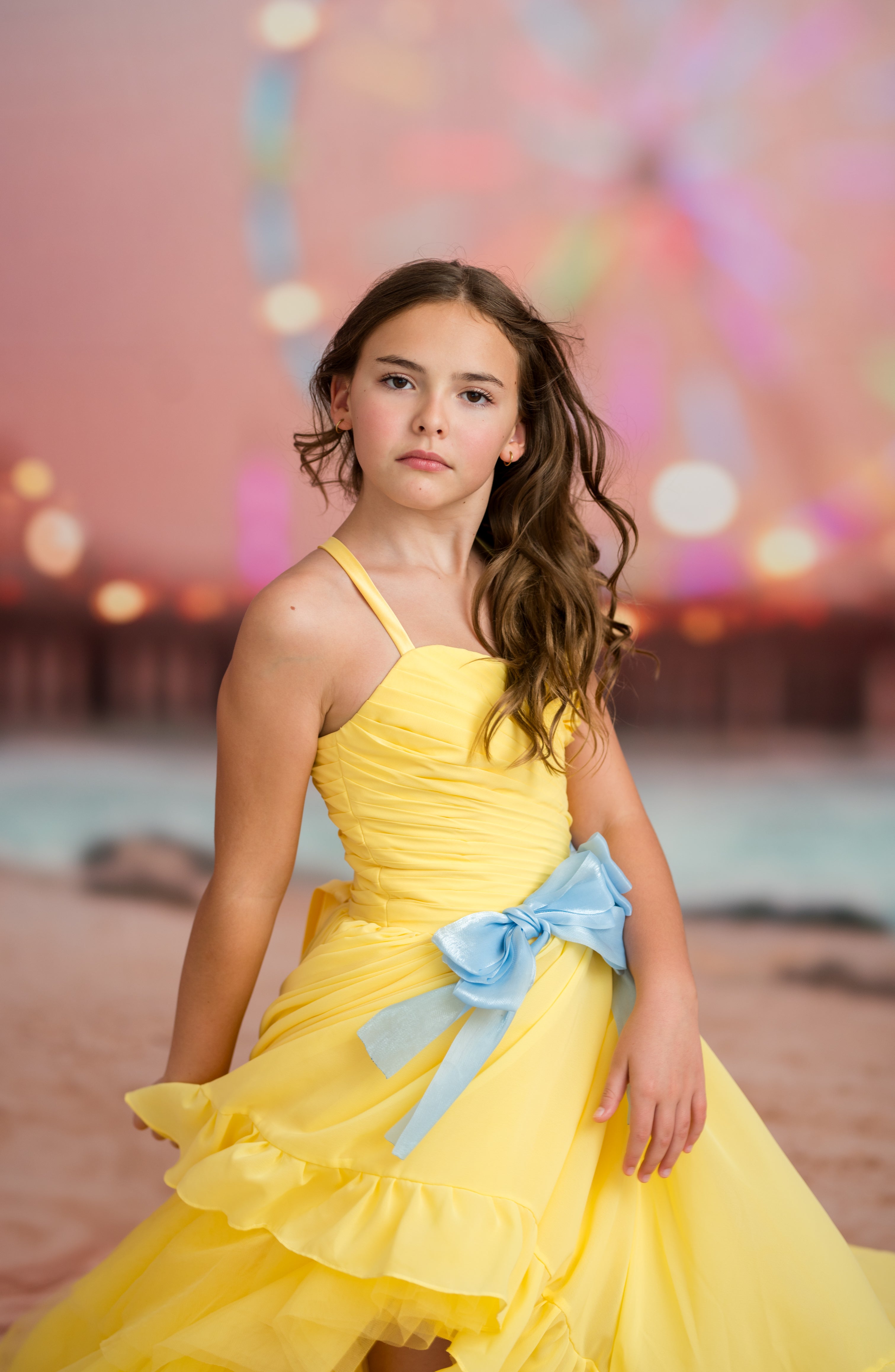 Young girl in a yellow dress with a blue bow standing on a beach at sunset.