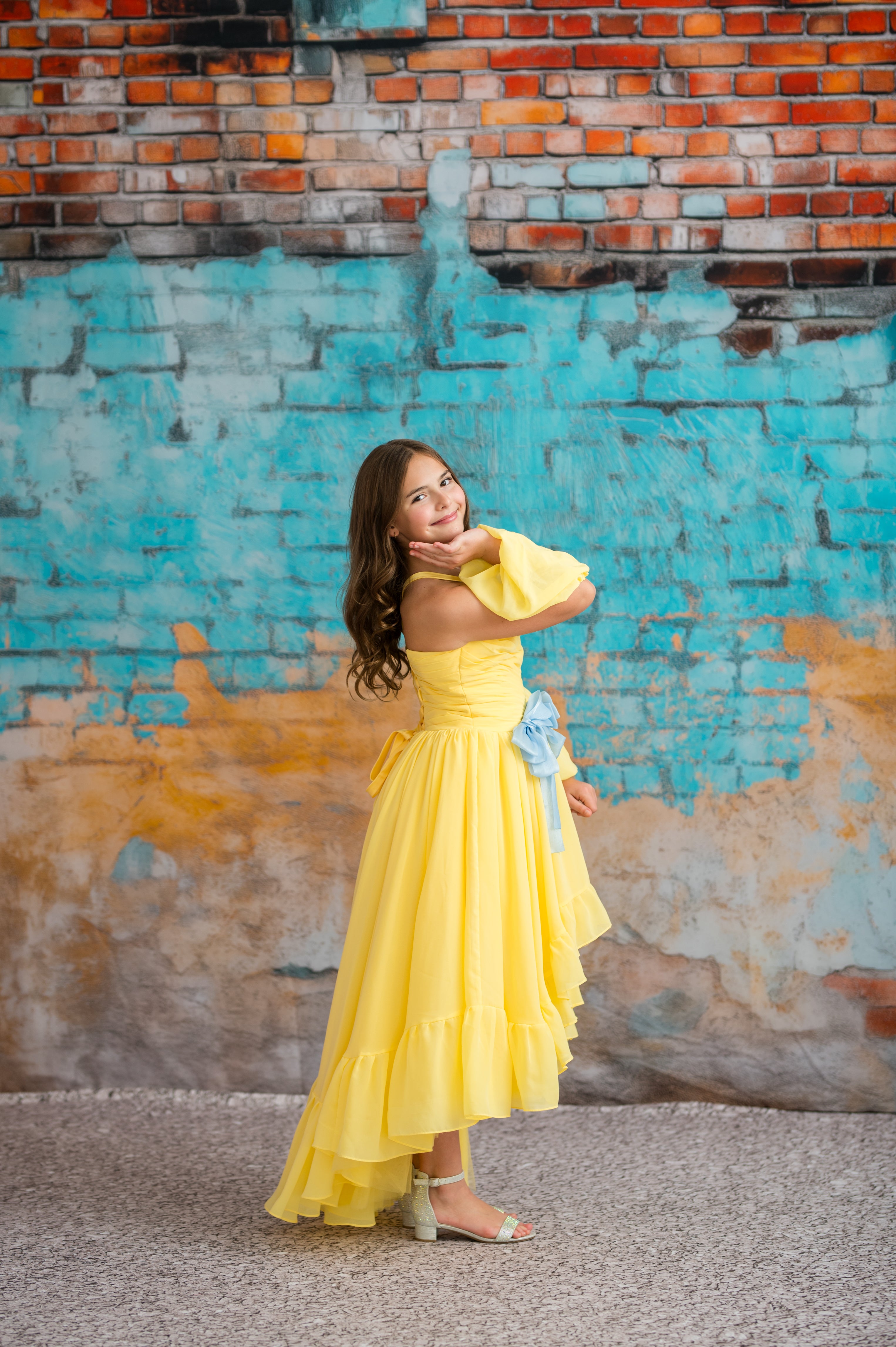 Woman in a yellow dress posing against a textured wall with blue and orange hues.