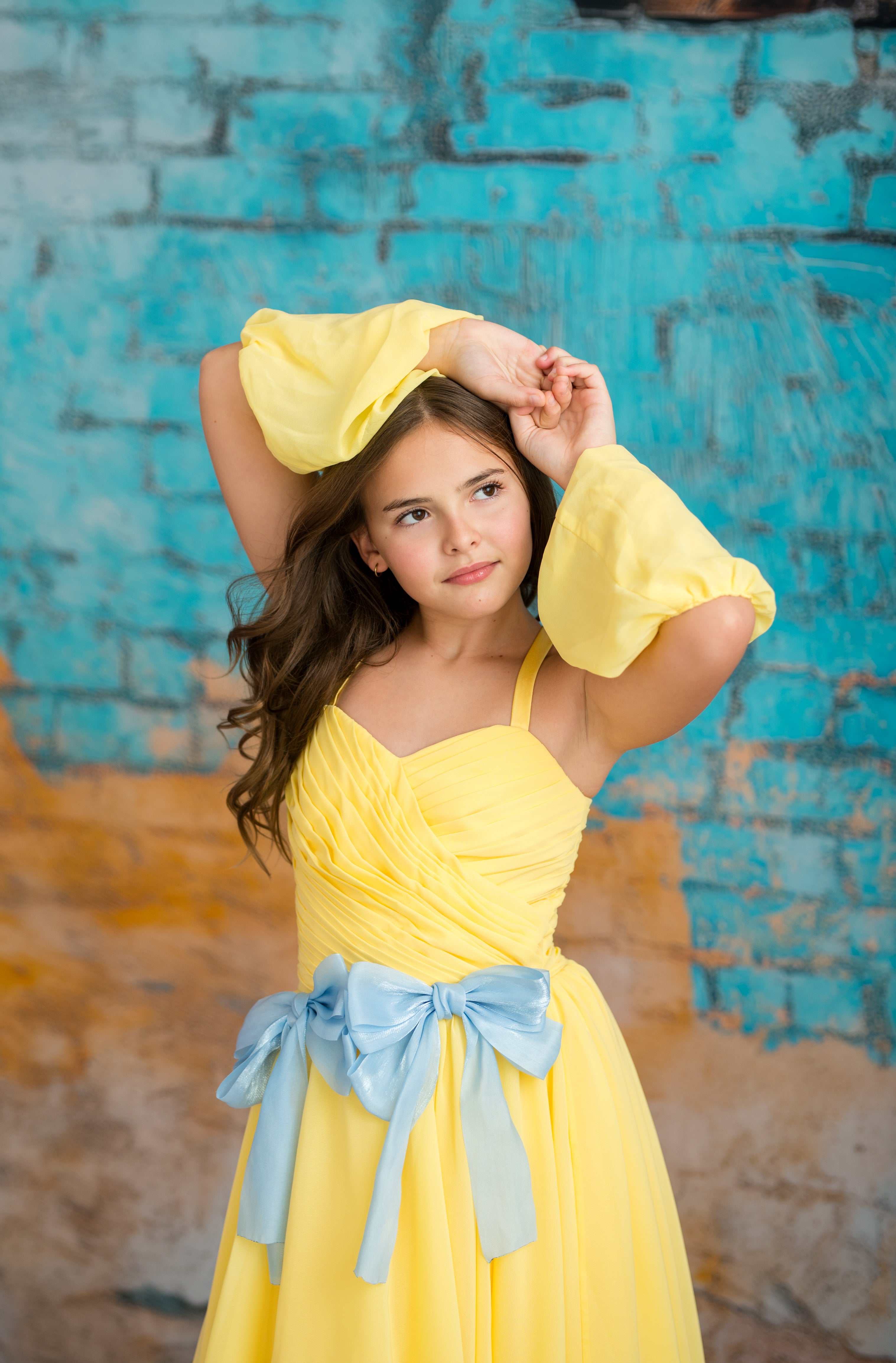 Young girl in a yellow dress with a blue sash against a textured blue and orange wall.