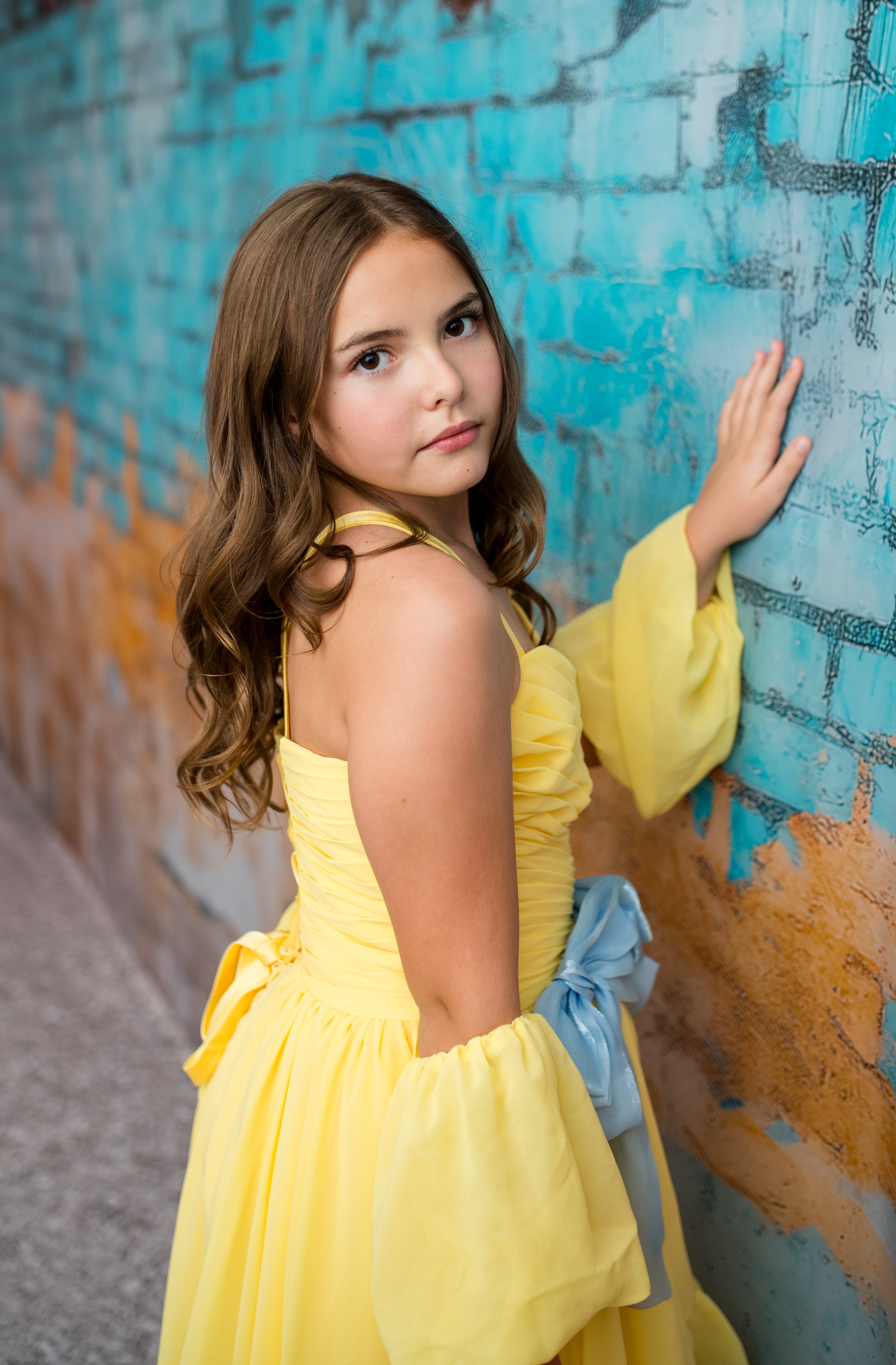 Young girl in a yellow dress standing against a textured blue and orange wall.