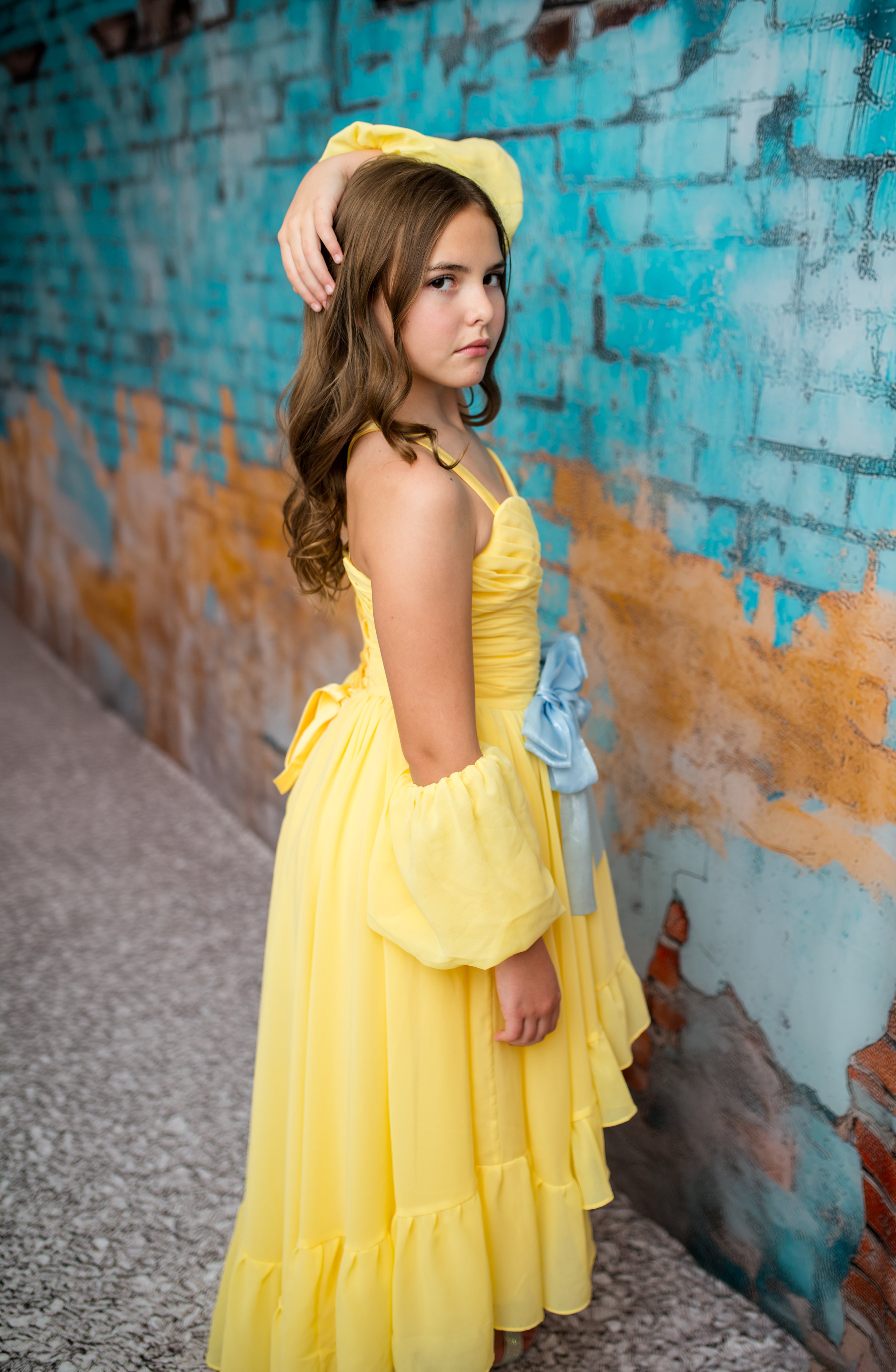 Young girl in a yellow dress standing against a textured blue wall.
