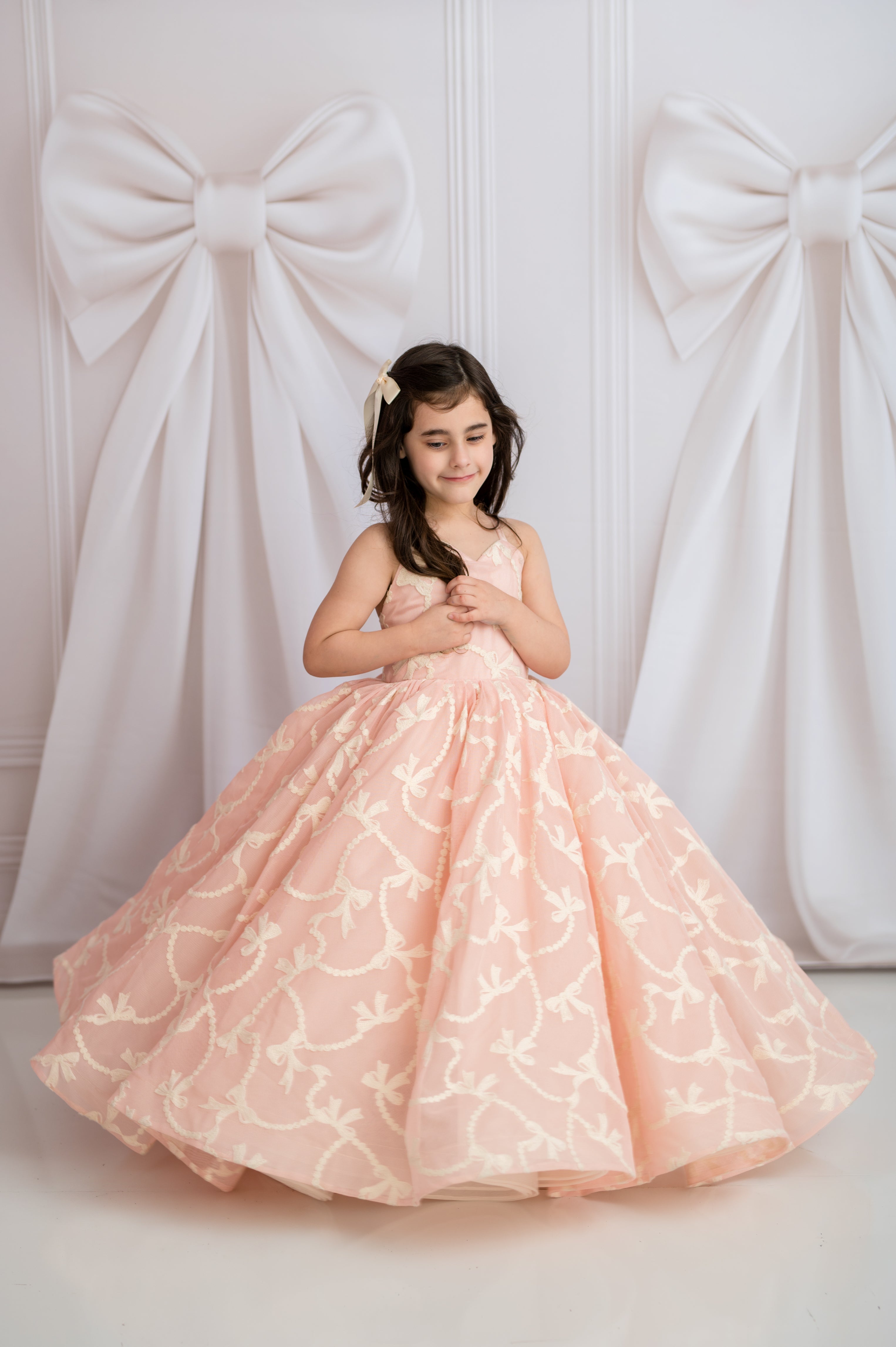 Young girl in a pink floral dress standing against a white wall with large decorative bows.