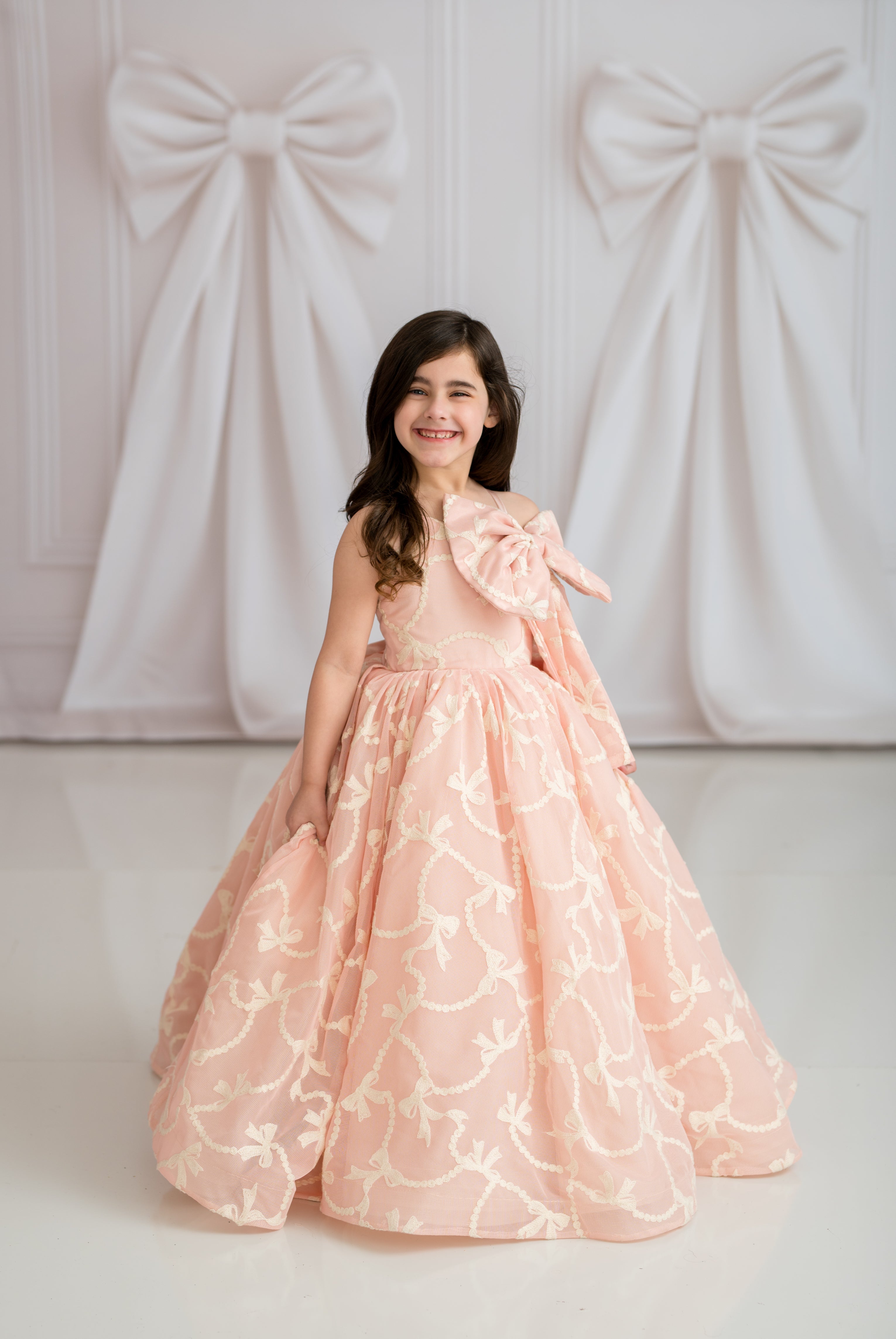 Young girl in a pink floral dress standing against a white wall with decorative bows.