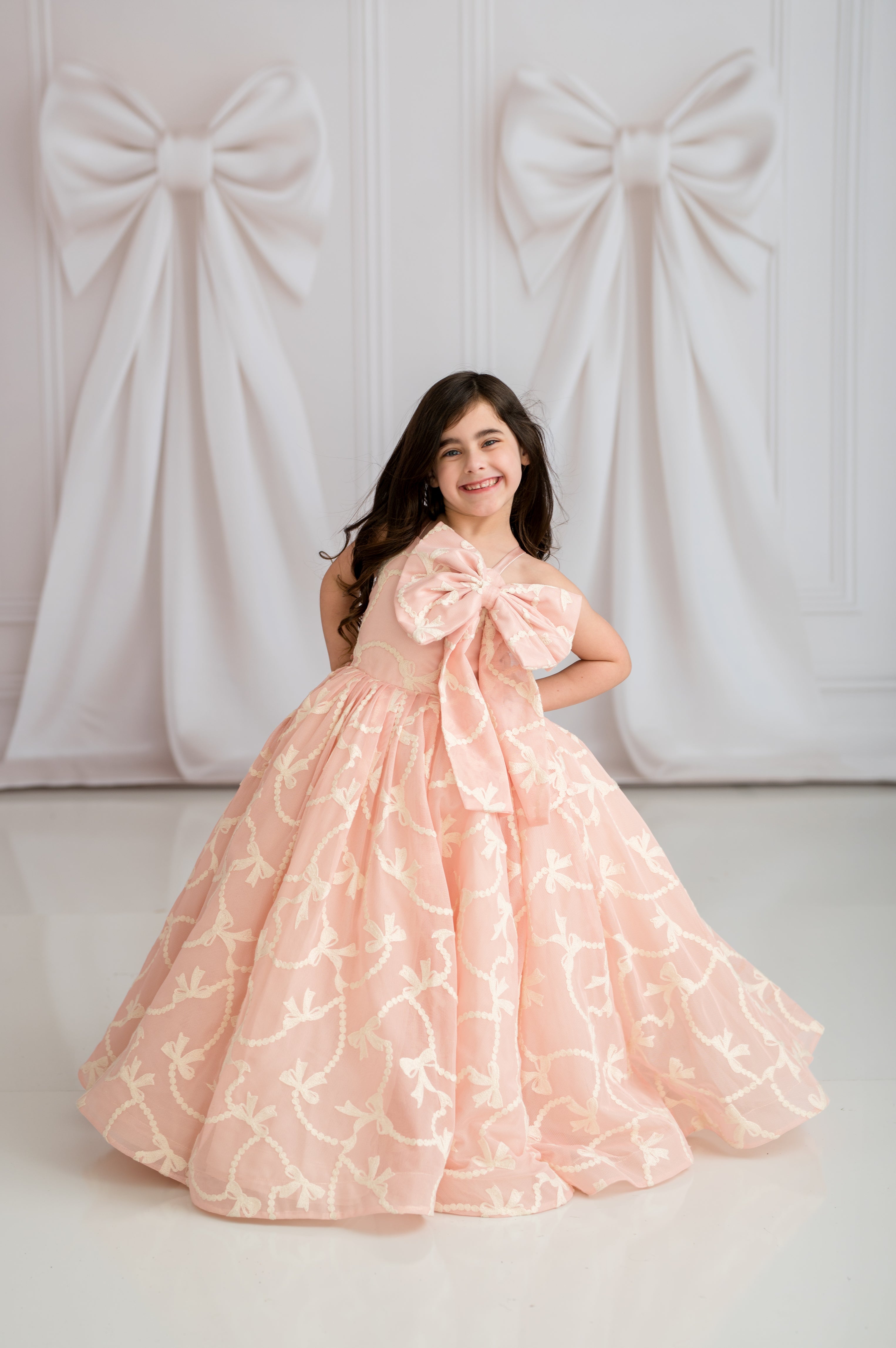 Young girl in a pink gown with a large bow standing against a white background with decorative bows.