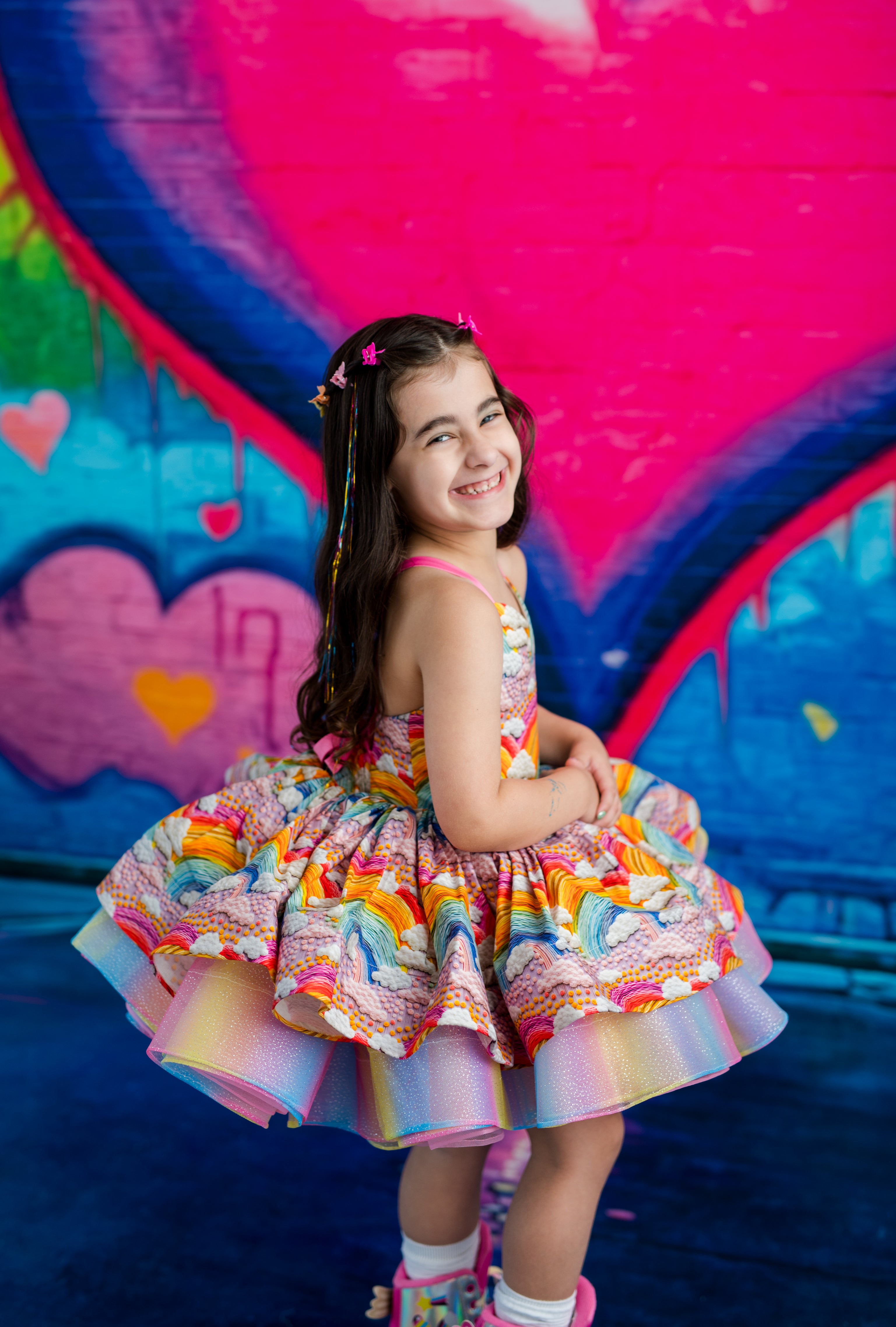 A young girl with long dark hair smiles while wearing a colorful, tiered ruffle dress with a rainbow and floral pattern, posed in front of a vibrant graffiti wall with large pink and blue heart shapes.